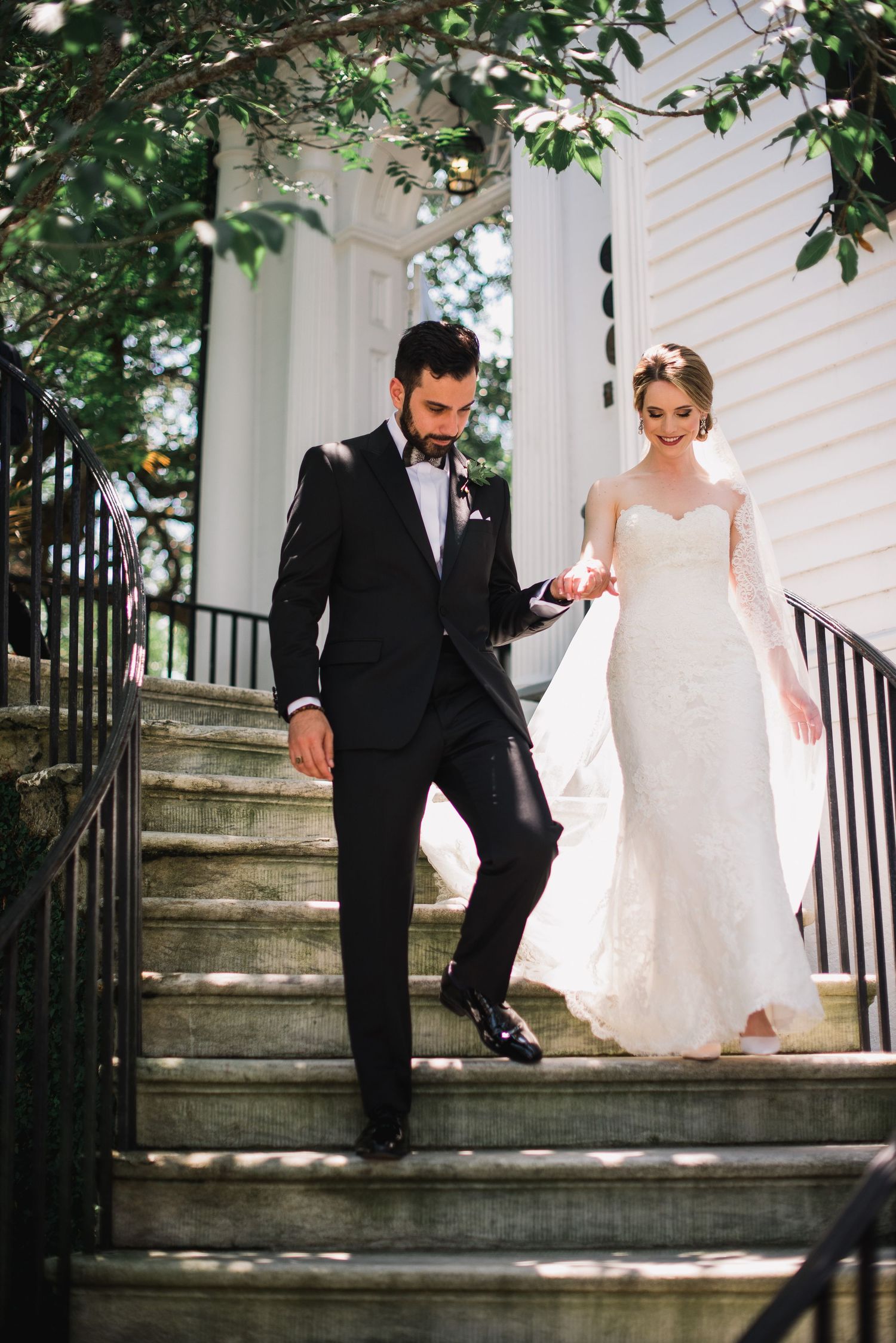 Newlyweds descend stone steps together outside a white building with greenery.