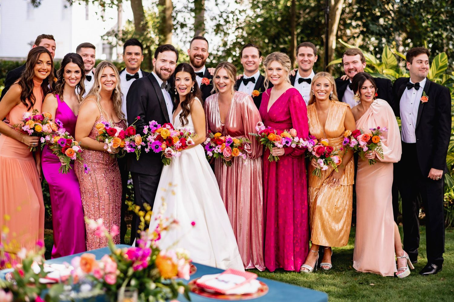 Wedding party members in colorful pink and coral dresses gather outdoors for celebratory group photos.