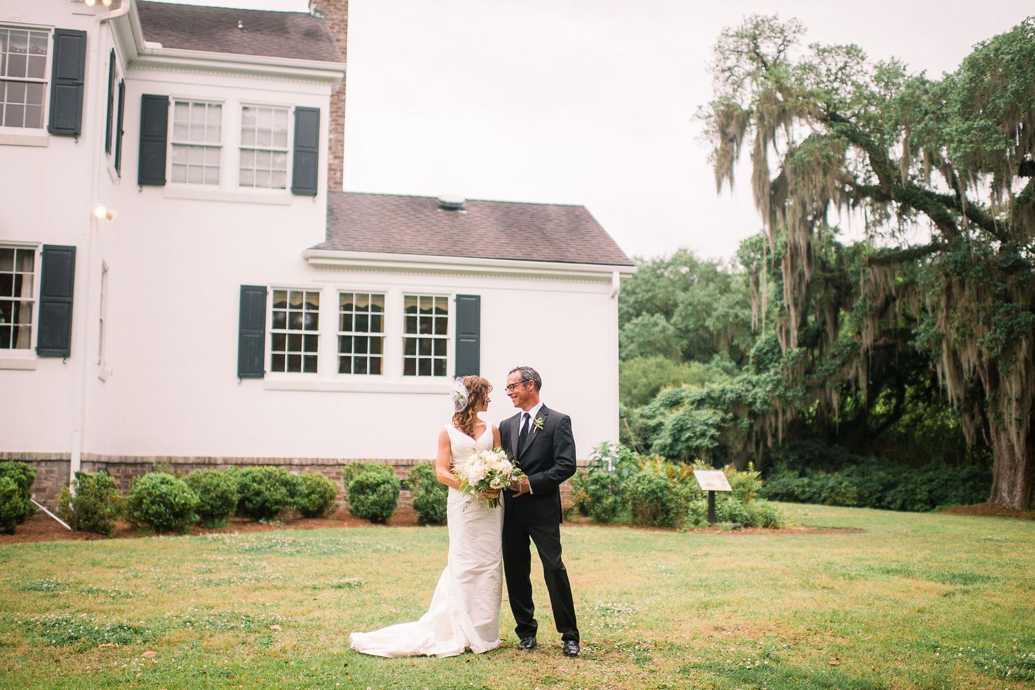 A couple shares an intimate moment on the lawn of a grand white plantation-style house with Spanish moss-draped trees.