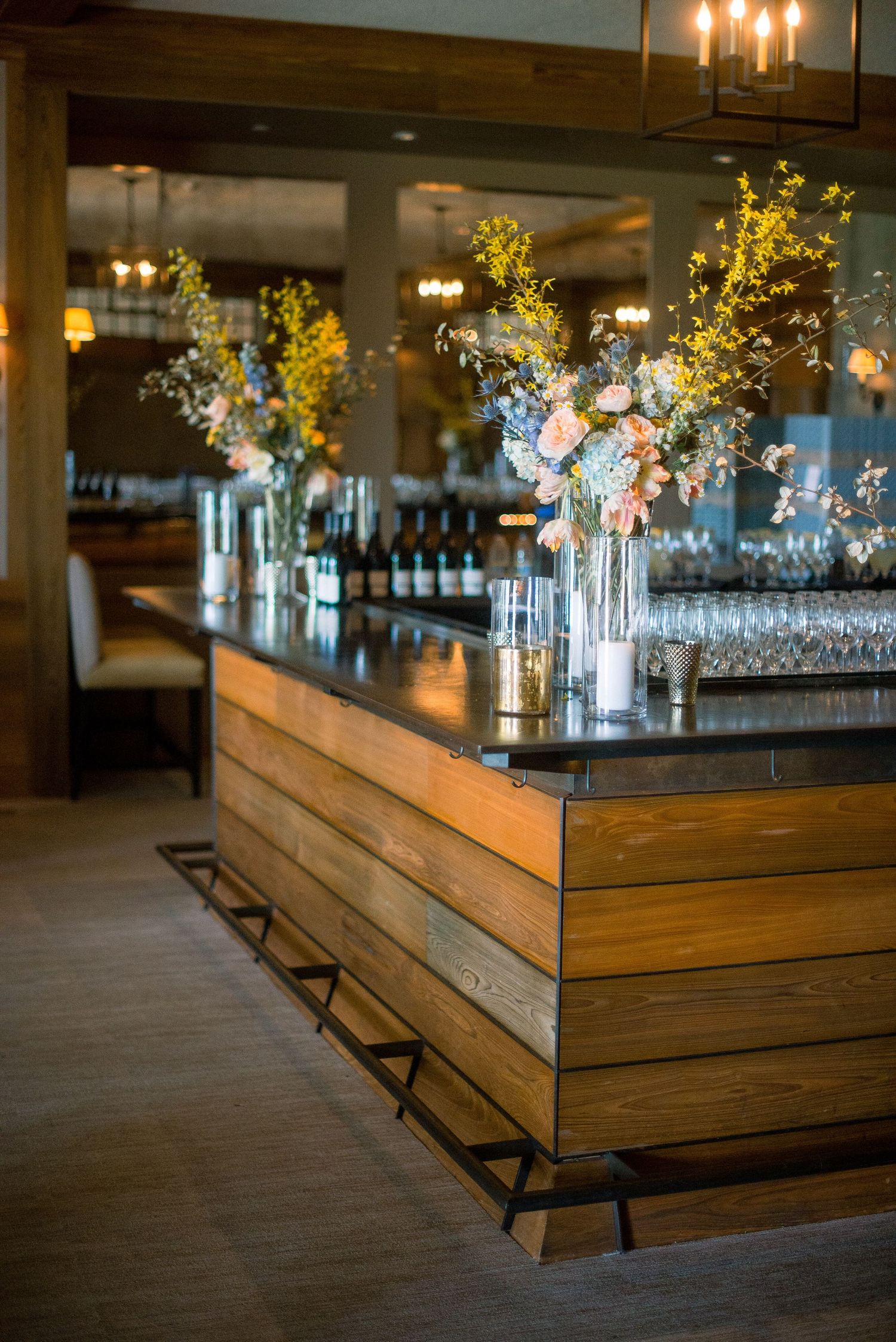 Elegant wooden bar counter with lit candles and floral centerpieces in glass vases along mirrored backsplash.