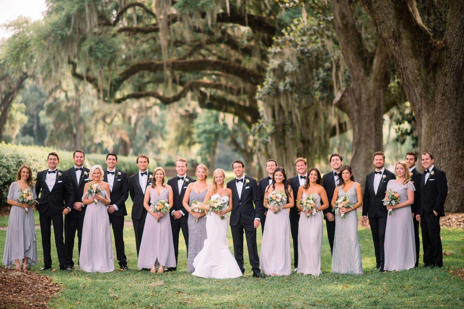 Wedding party in formal attire poses under Spanish moss draped oak trees at an elegant outdoor venue.