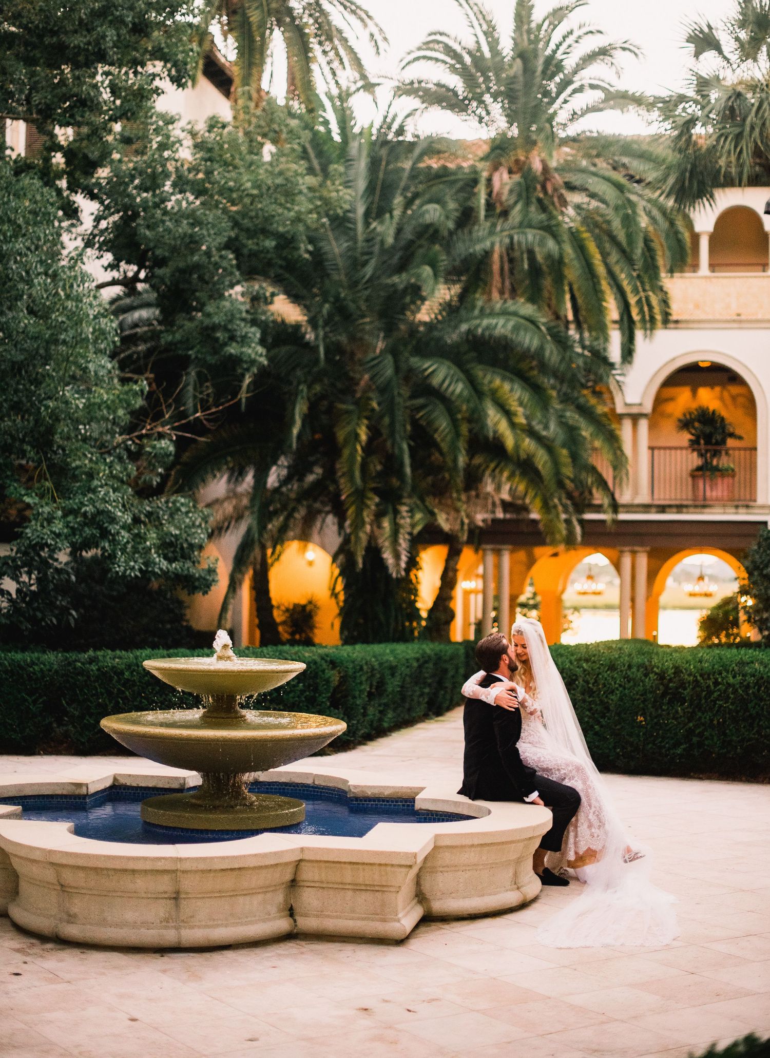 A couple shares a romantic moment by an ornate stone fountain surrounded by palm trees in an elegant courtyard at dusk.