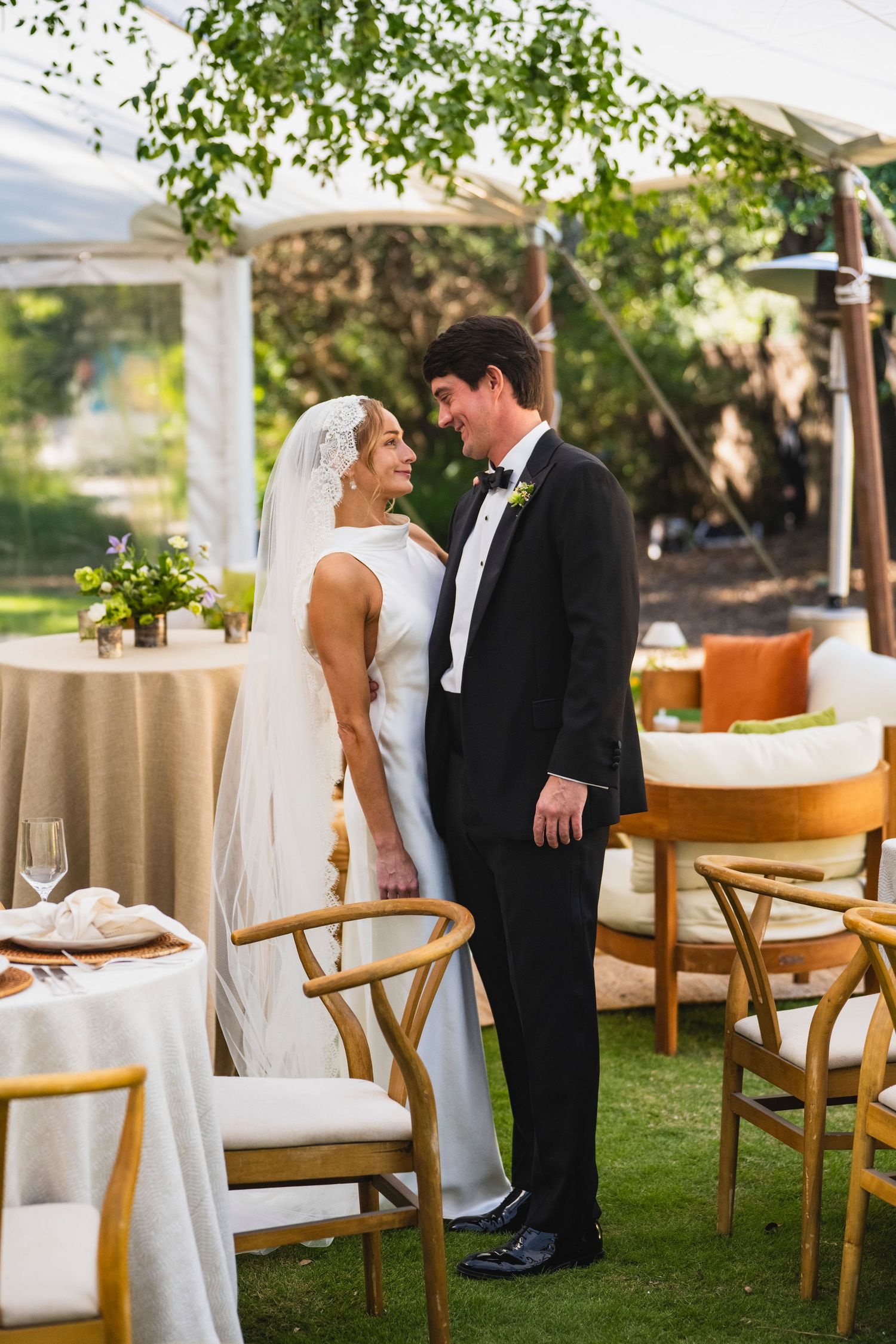 A couple in formal wedding attire shares an intimate moment in a garden reception setting with wooden chairs and white decor.
