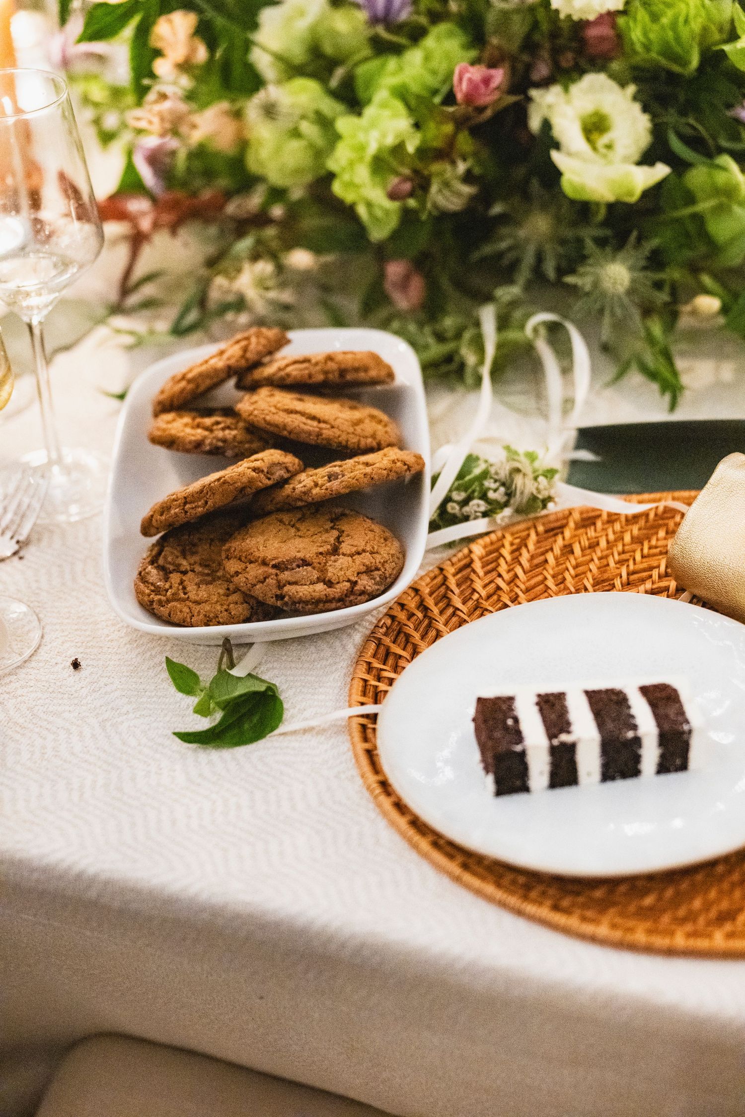 Elegant table setting with chocolate cookies and desserts on white plates with floral decorations and woven placemats.