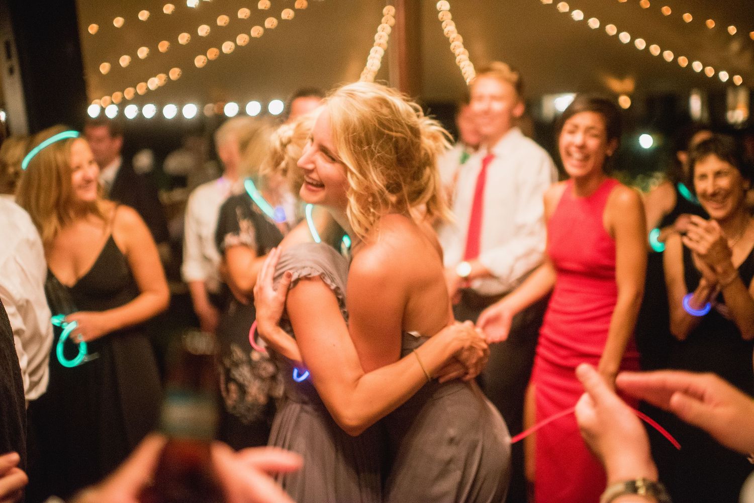 People dancing and celebrating at a wedding reception under glowing string lights at night.