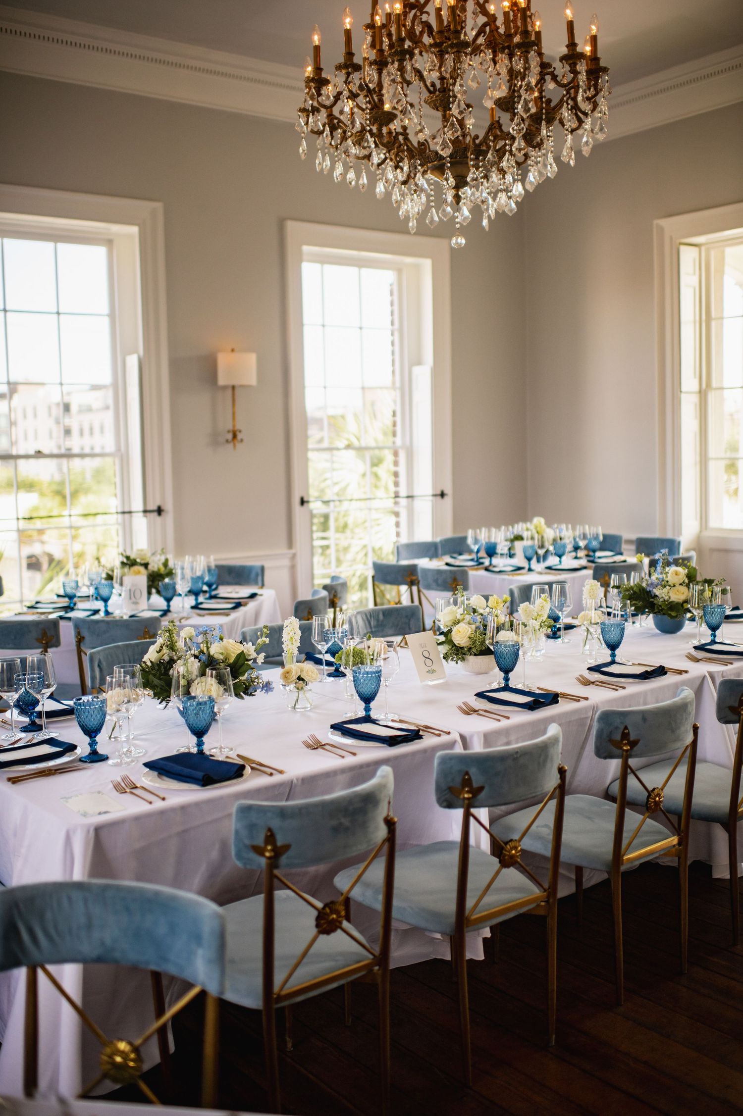 Elegant dining room with crystal chandelier, white tablecloth, blue accents, and tall windows overlooking a scenic view.