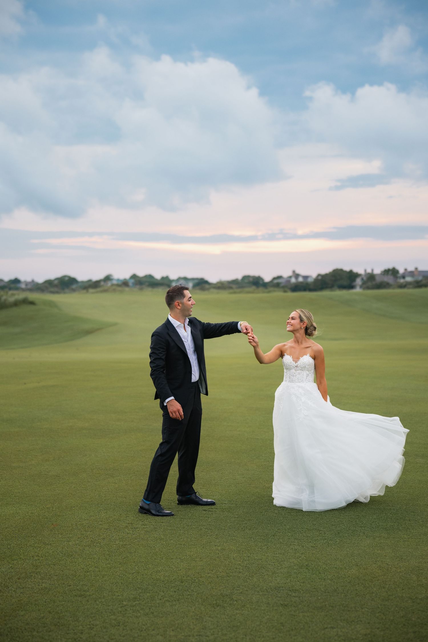 Couple in wedding attire playfully dancing and twirling on a vast green golf course under blue skies.