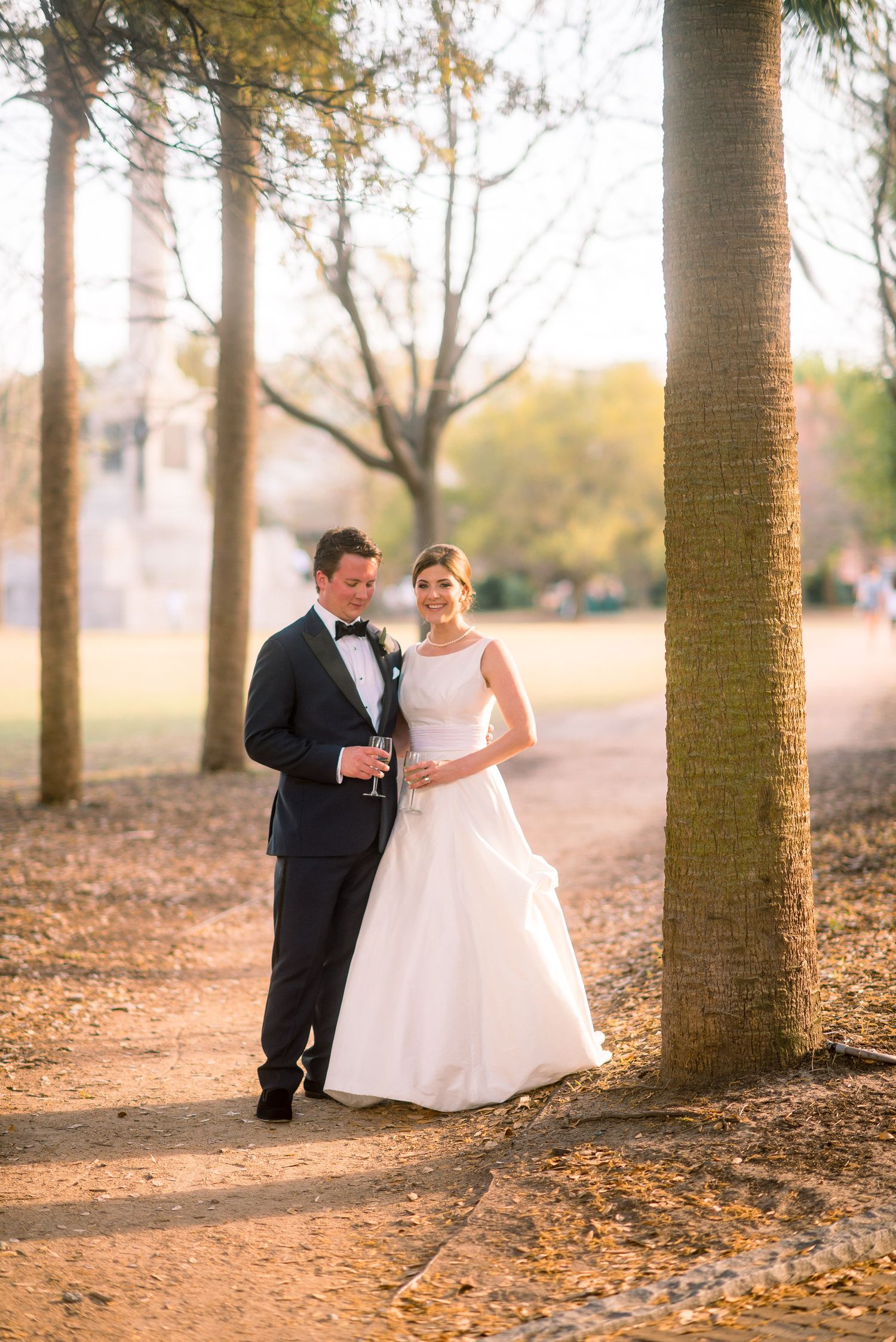 A couple in formal wedding attire pose together on a dirt path among tall trees during golden hour.