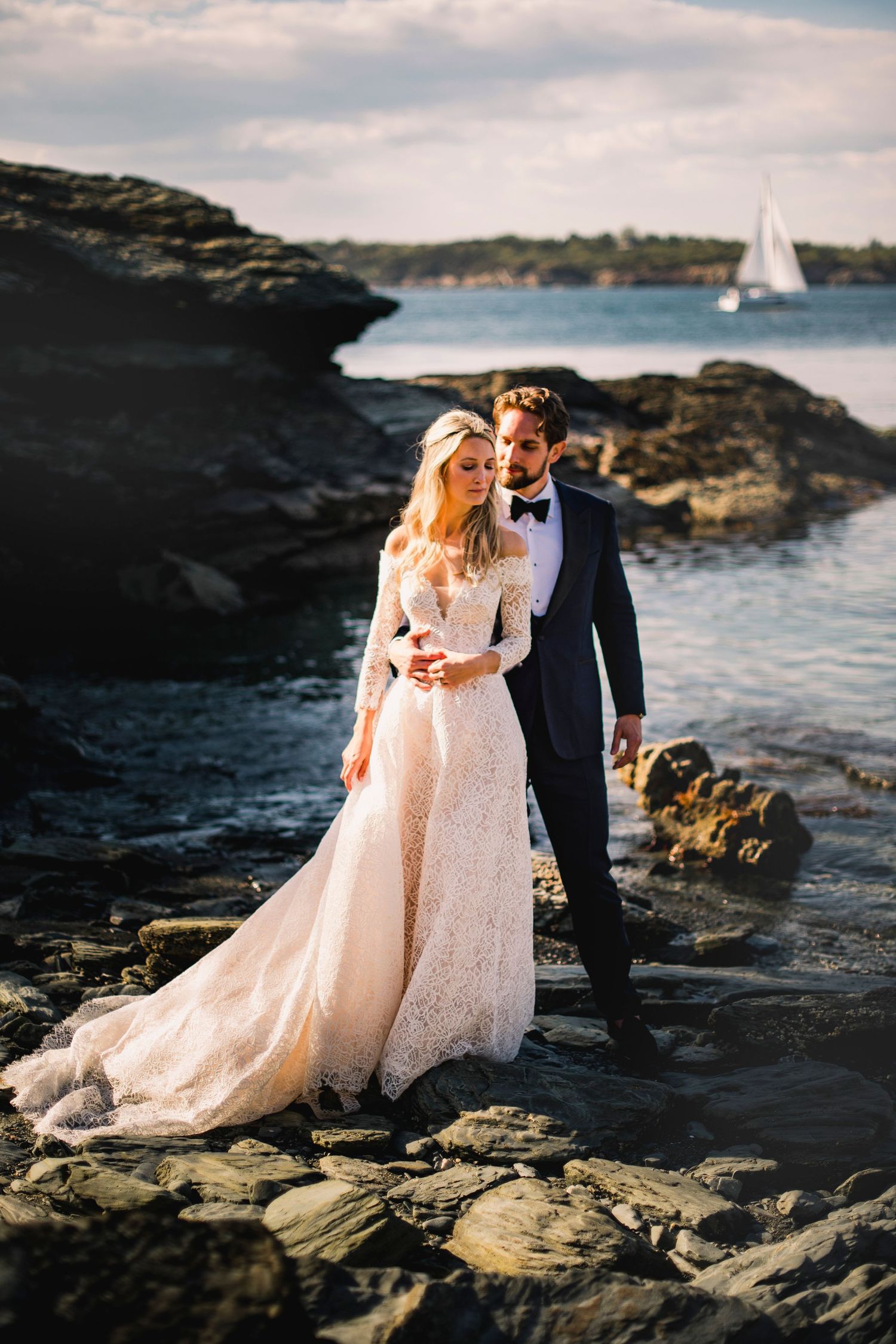 A romantic coastal wedding photoshoot featuring a couple on rocky cliffs with a sailboat in the distance.