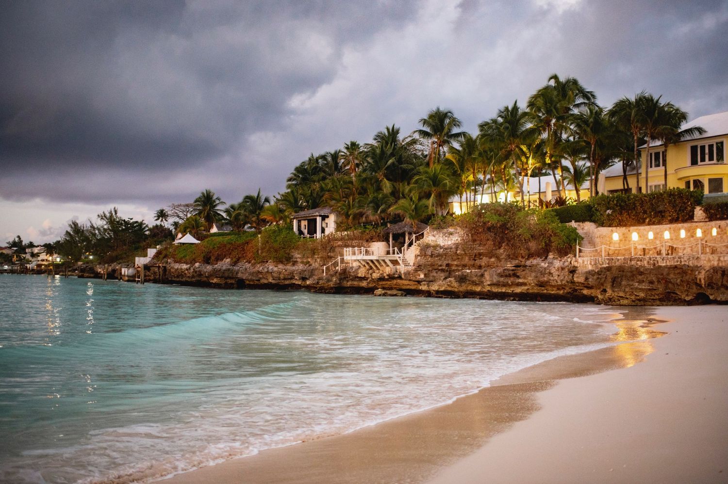 Luxurious beachfront homes line a tropical shore with turquoise waters and dramatic stormy skies.