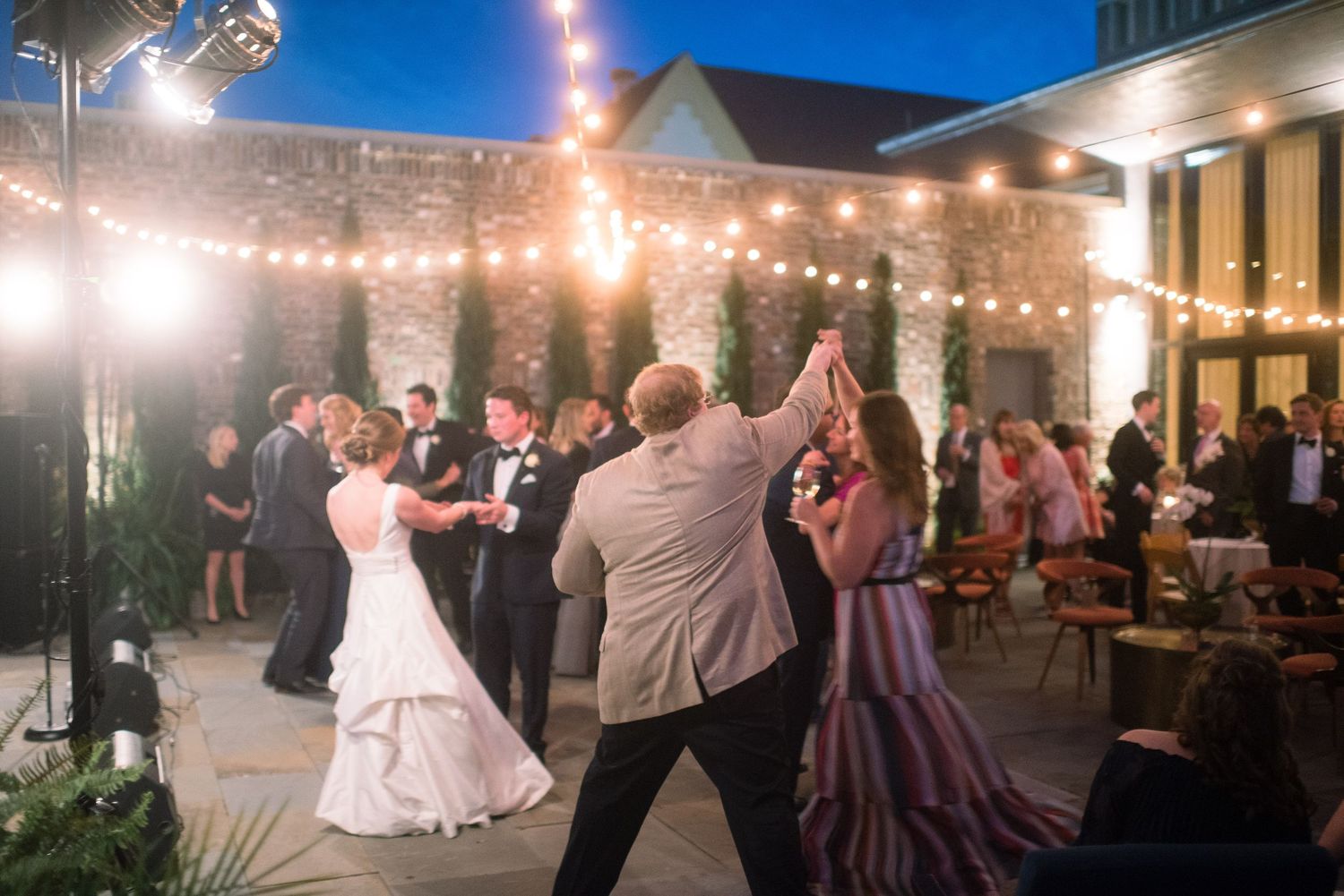 Wedding reception guests dancing and celebrating under string lights at an outdoor evening venue.