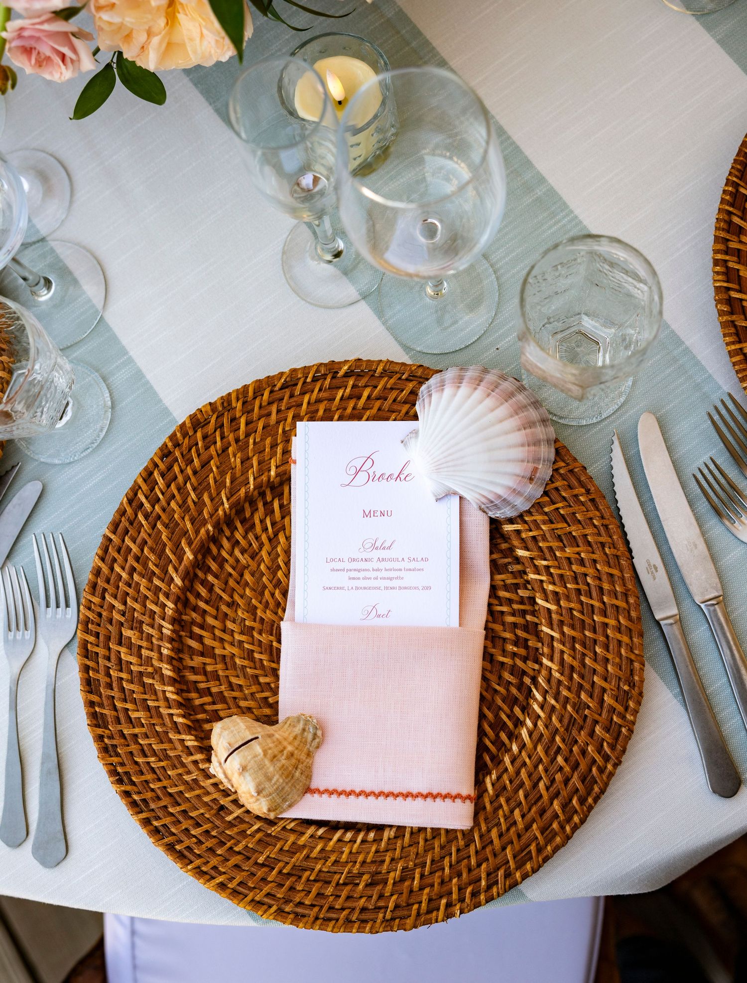 Coastal-themed place setting with seashell, woven charger plate, and pink menu card.