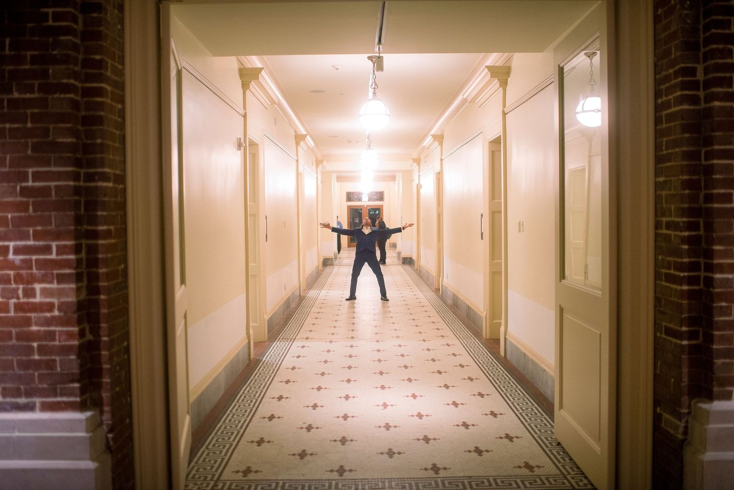 A figure stands at the end of a long, empty hallway with cream walls and tiled floors illuminated by overhead lighting.