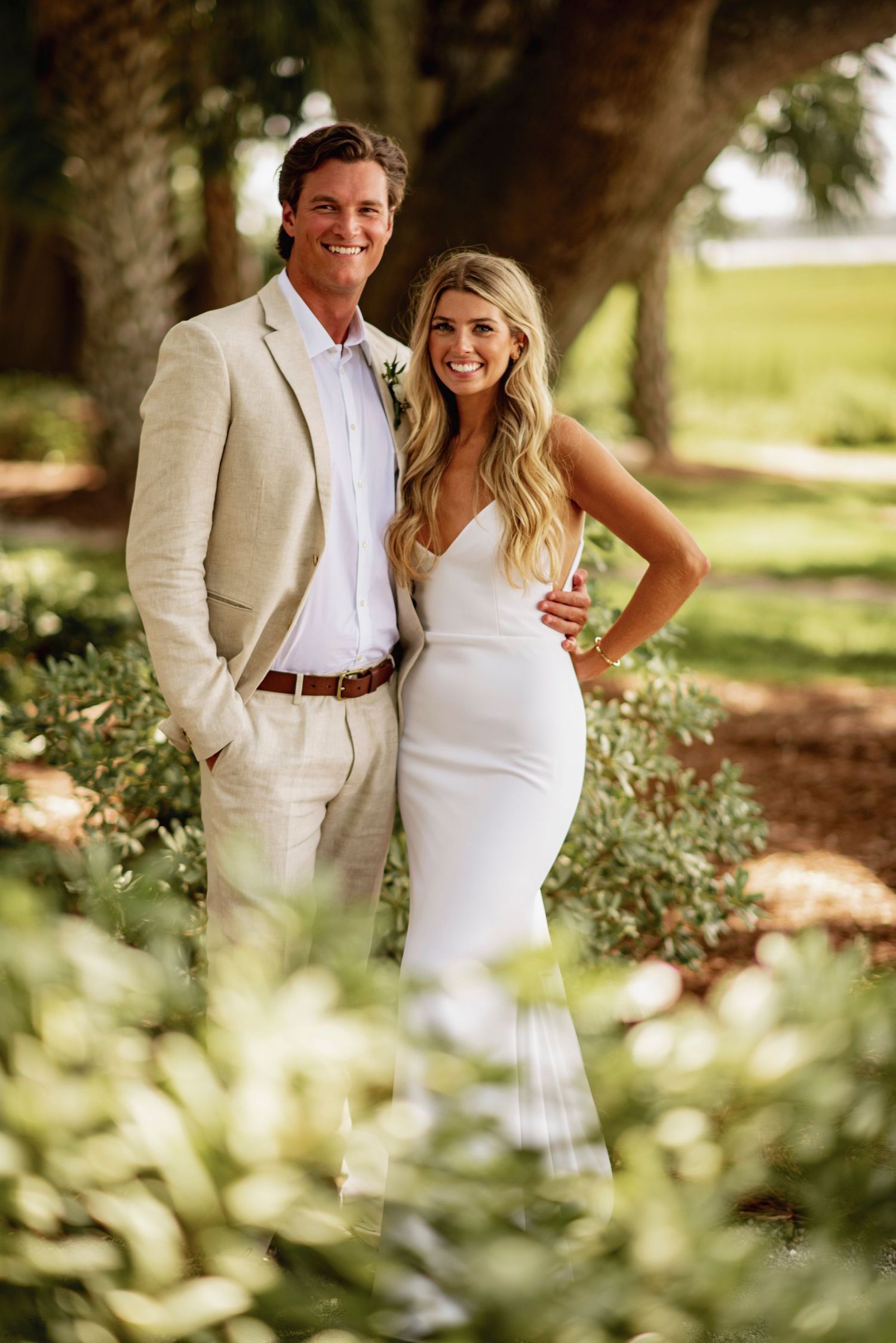 A couple in elegant white wedding attire poses together in a lush garden setting with soft natural lighting.
