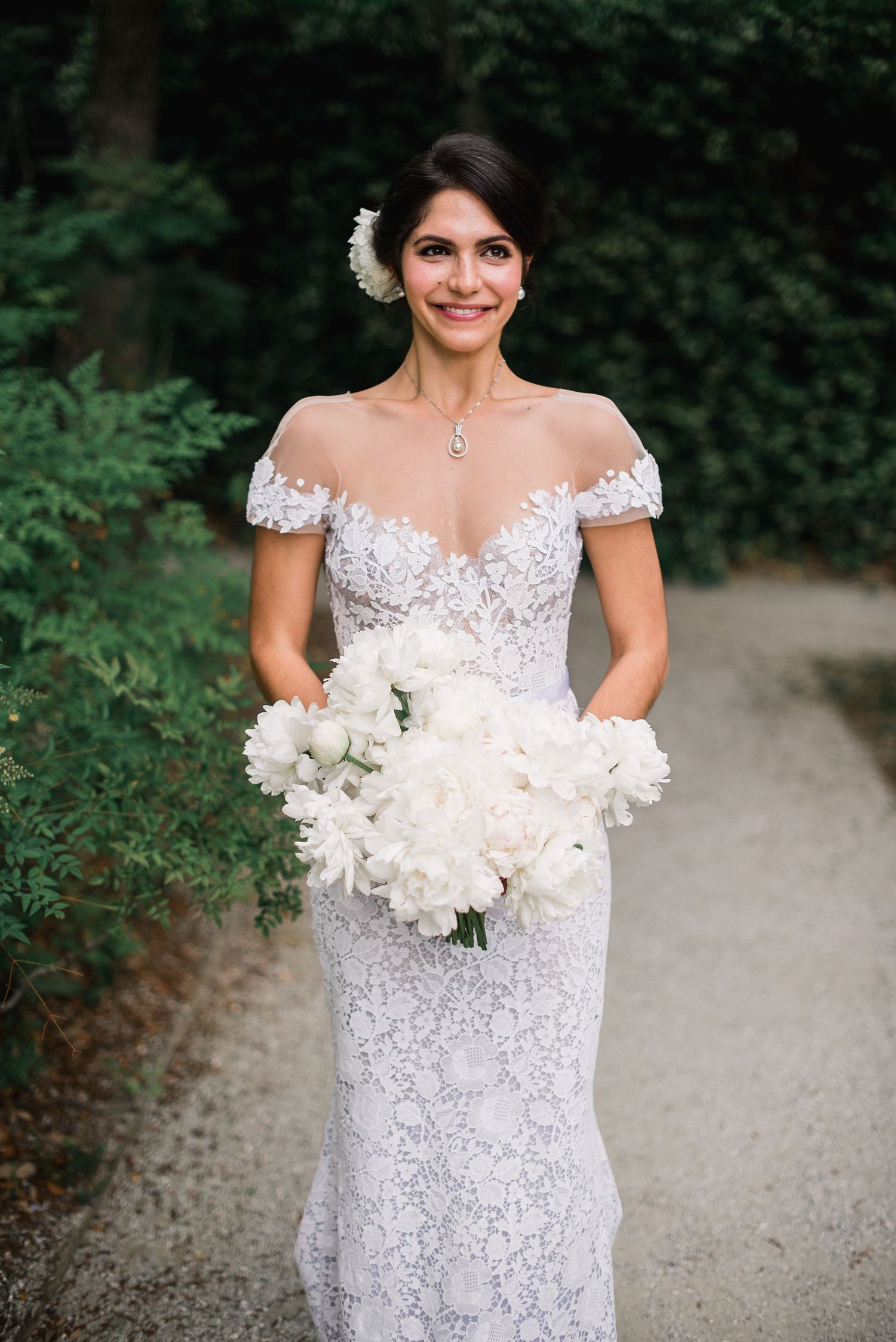 Bride in off-shoulder lace wedding gown holds elegant white peony bouquet while standing on garden path.