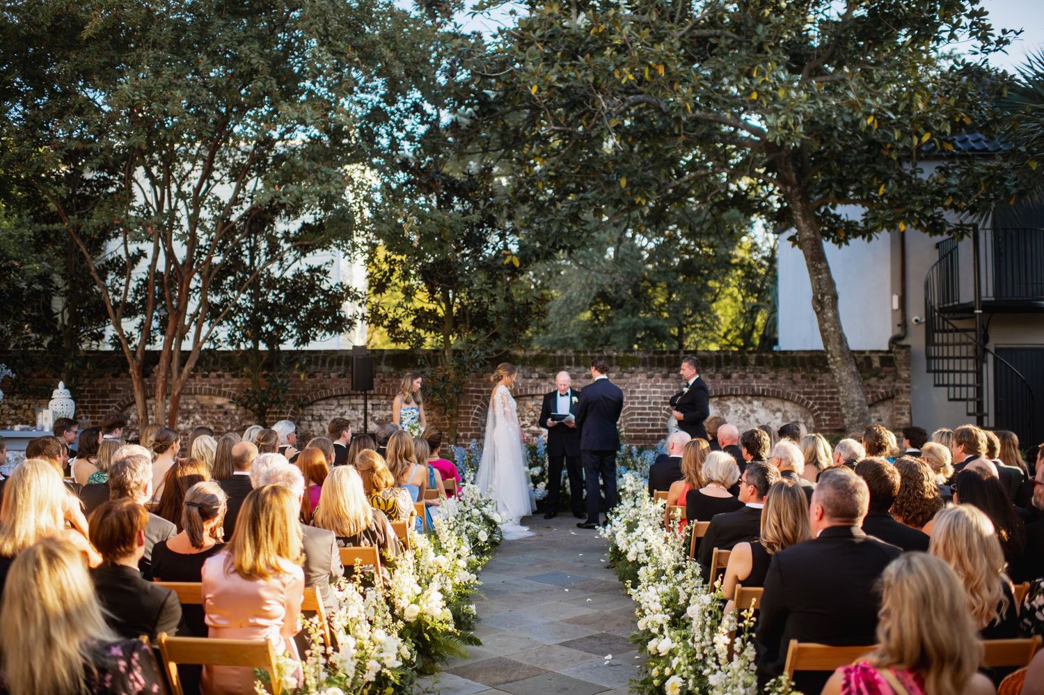 Wedding guests seated outdoors in rows of white chairs during a garden ceremony with floral decorations.