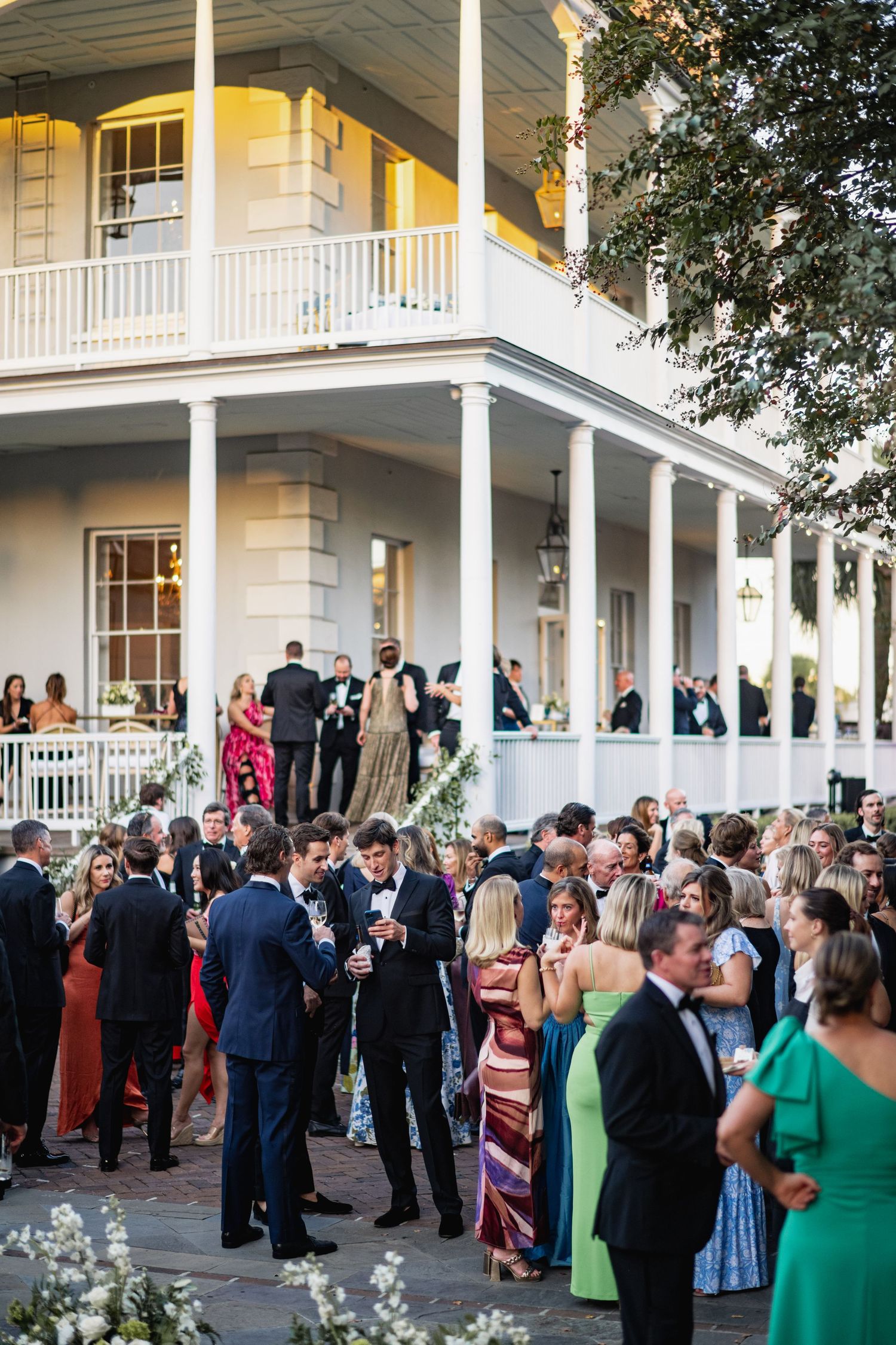 Wedding guests gather on the porch of a white colonial-style mansion during an elegant outdoor reception.