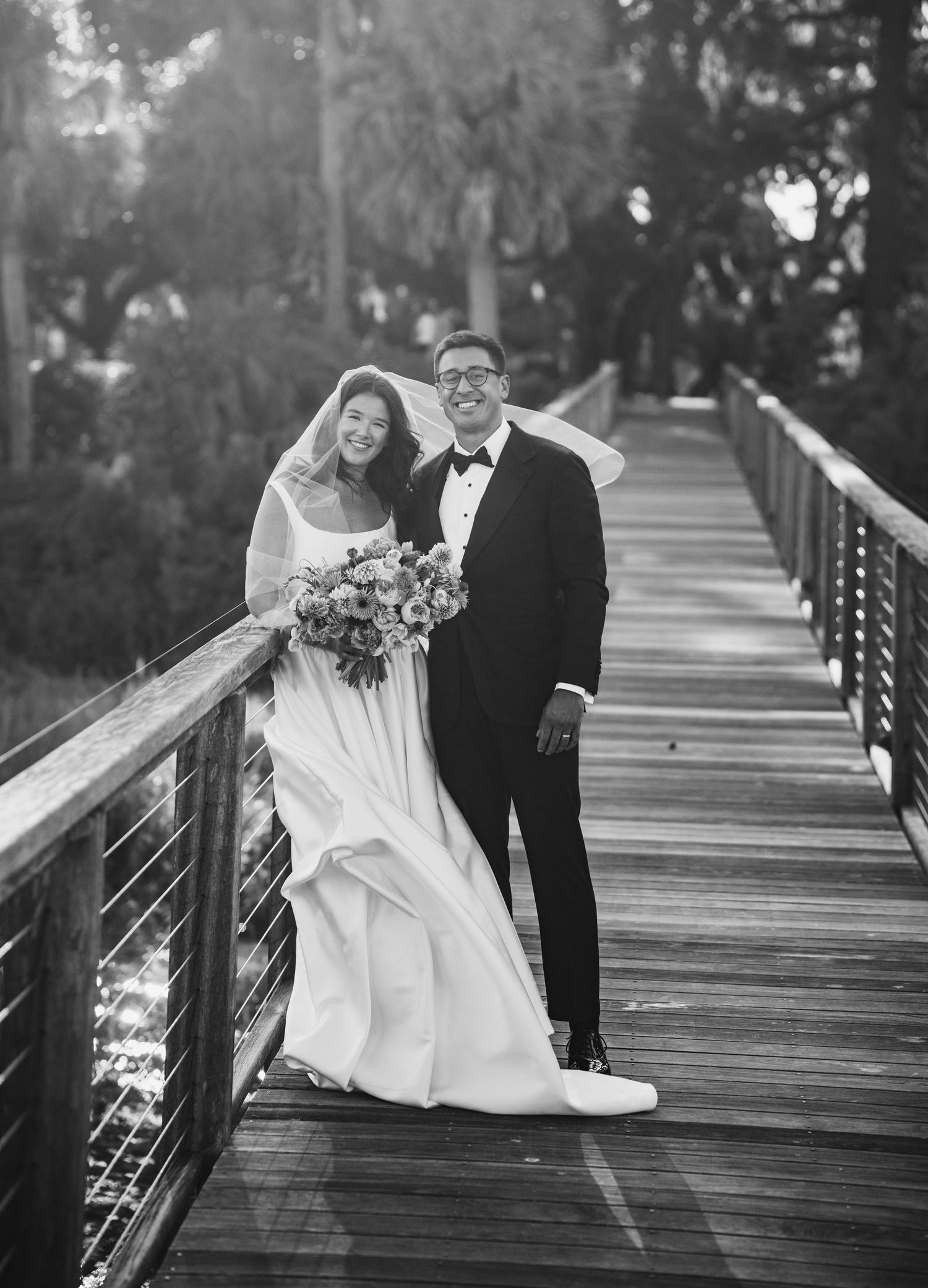 Black and white photo of a joyful wedding couple sharing a romantic moment on a wooden bridge surrounded by trees.