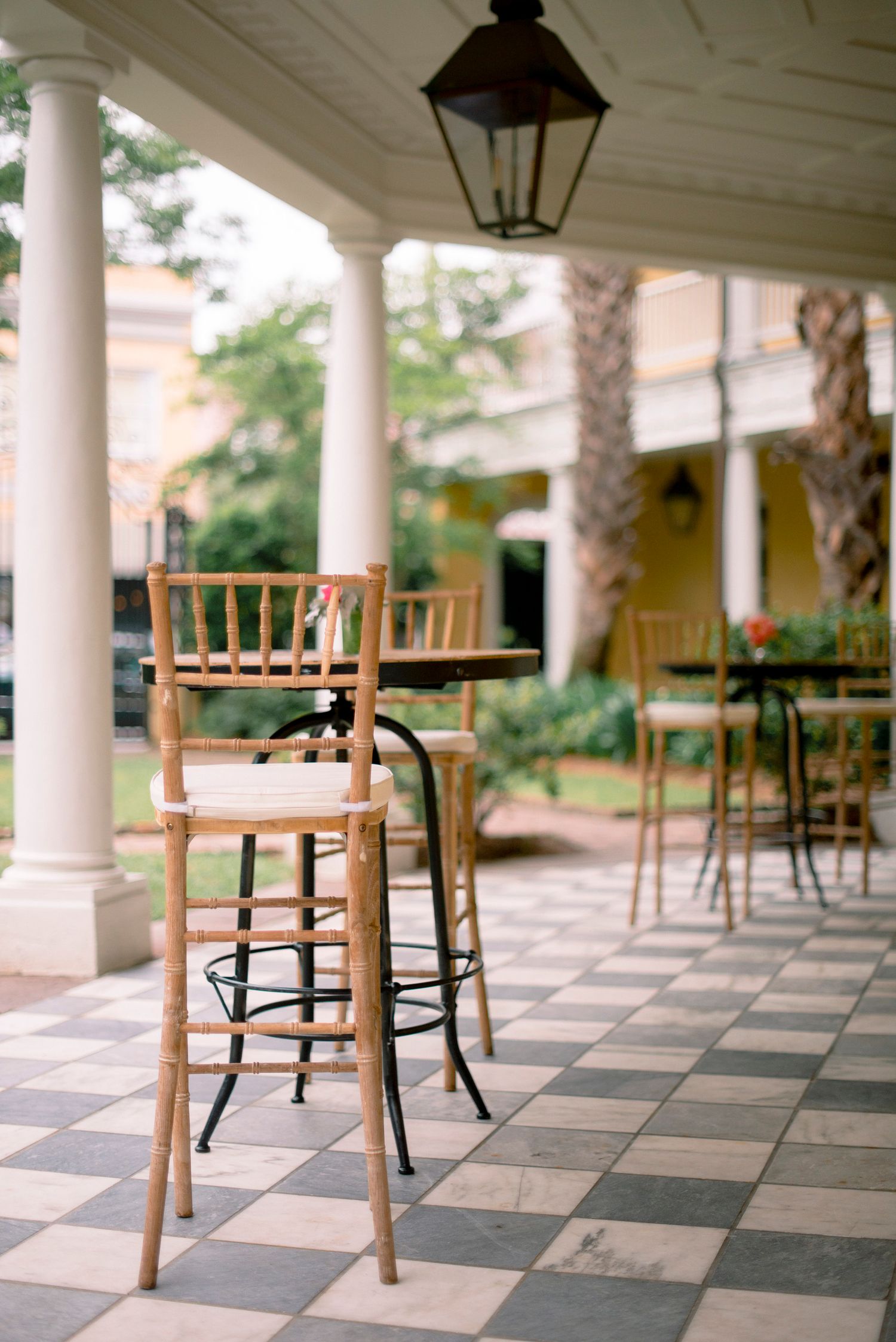 Empty wooden chairs line a covered porch with checkerboard tile flooring and white columns at a Southern estate.