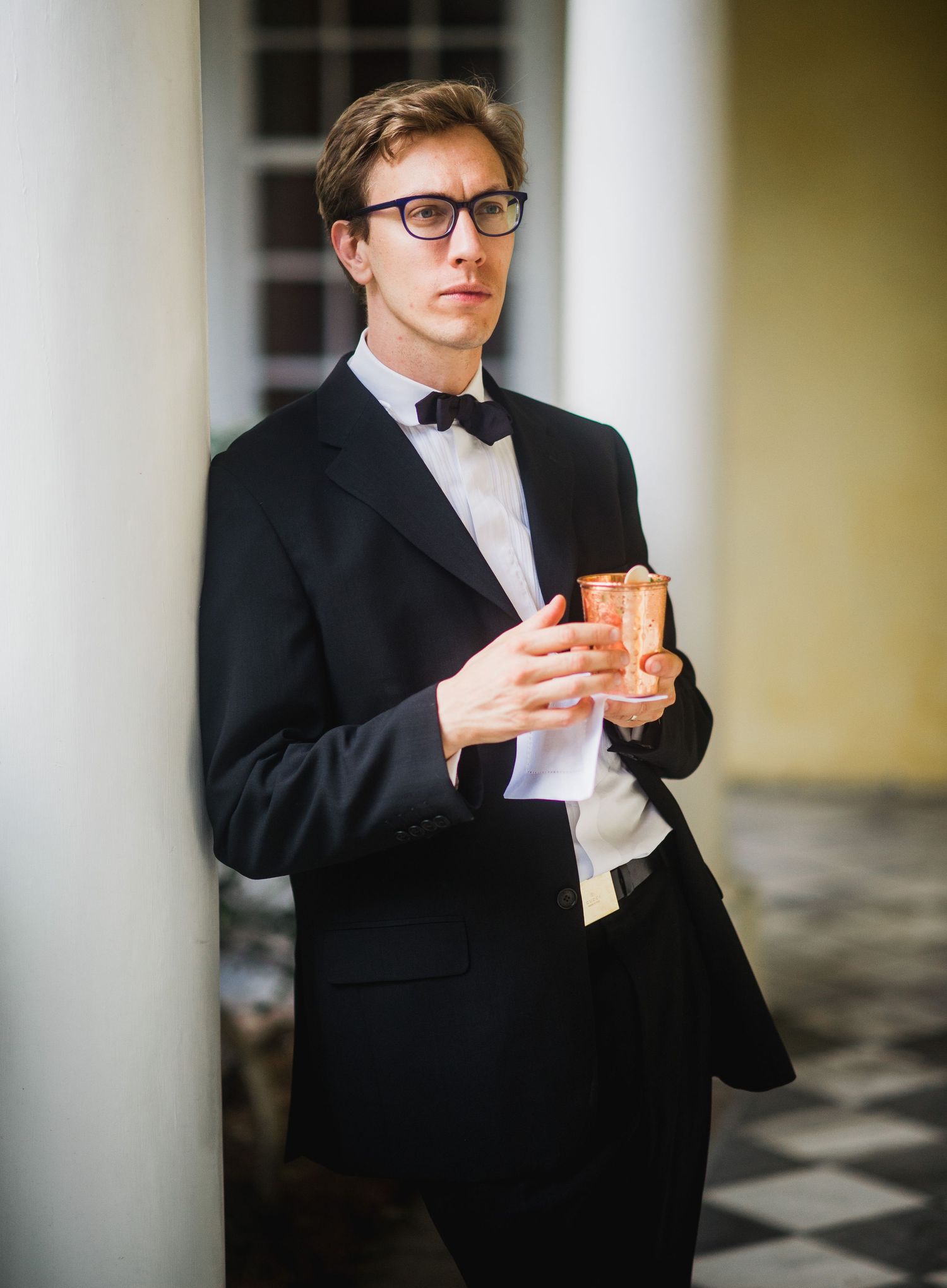 A person in a black suit and glasses holds a cup while standing in a room with checkered flooring.