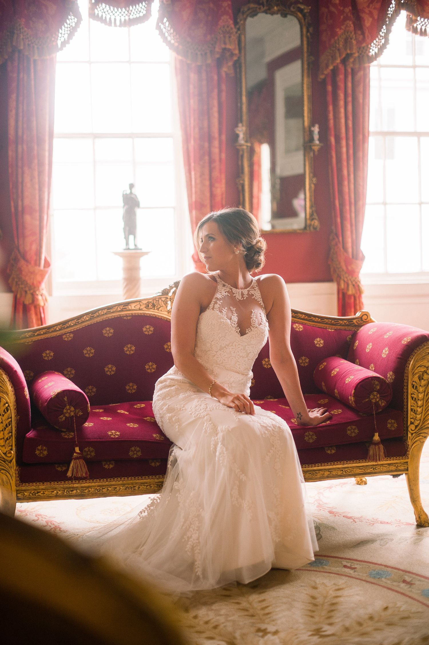 Elegant bride in lace gown sits on luxurious red Victorian sofa near sunlit windows in ornate room.