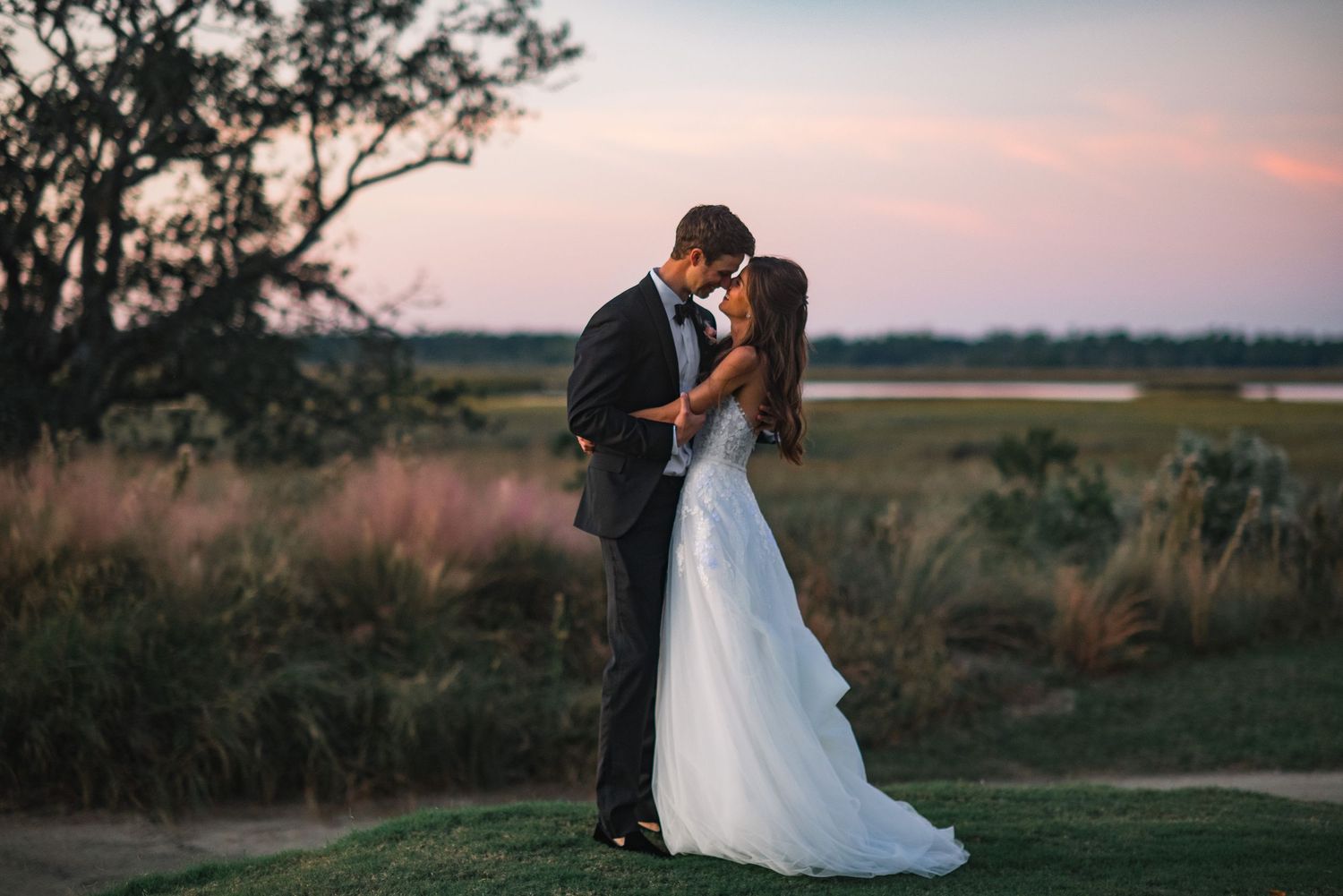 A couple shares a romantic kiss at sunset in a rustic outdoor setting with tall grass and trees in the background.