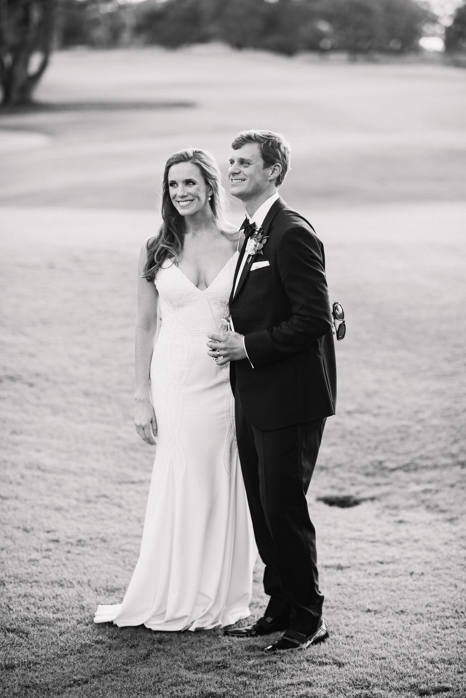 A black and white wedding photo series shows a couple standing together on a grassy lawn.