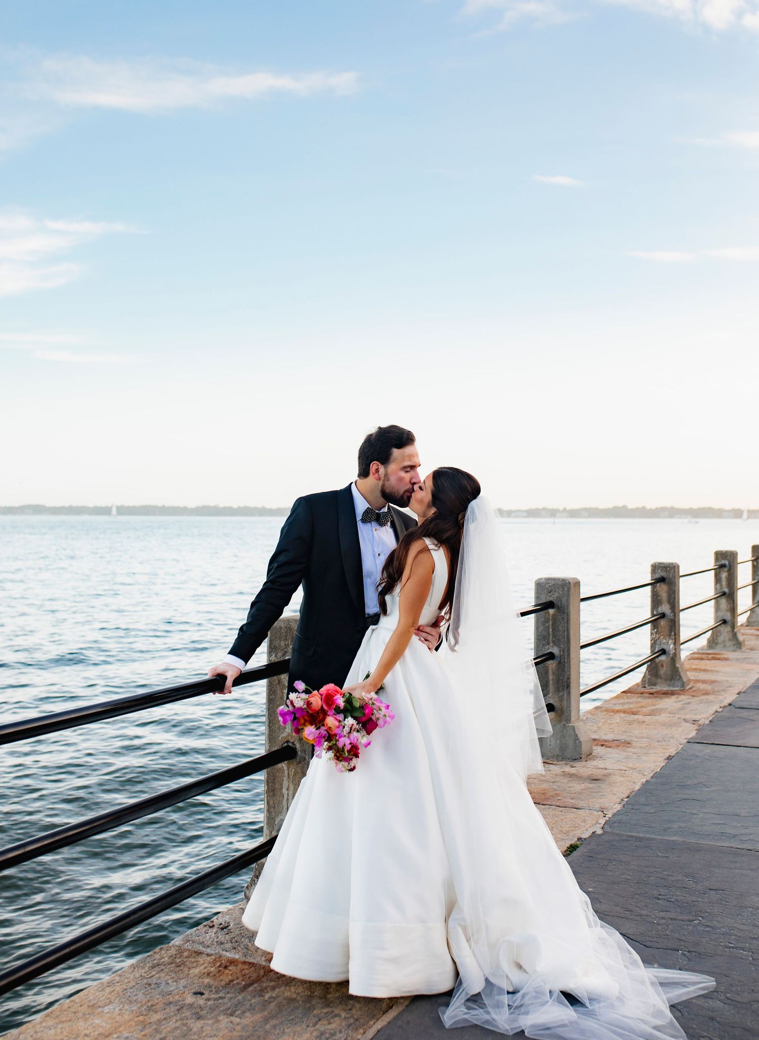 A couple in wedding attire shares romantic moments on a wooden pier overlooking a serene waterfront at sunset.