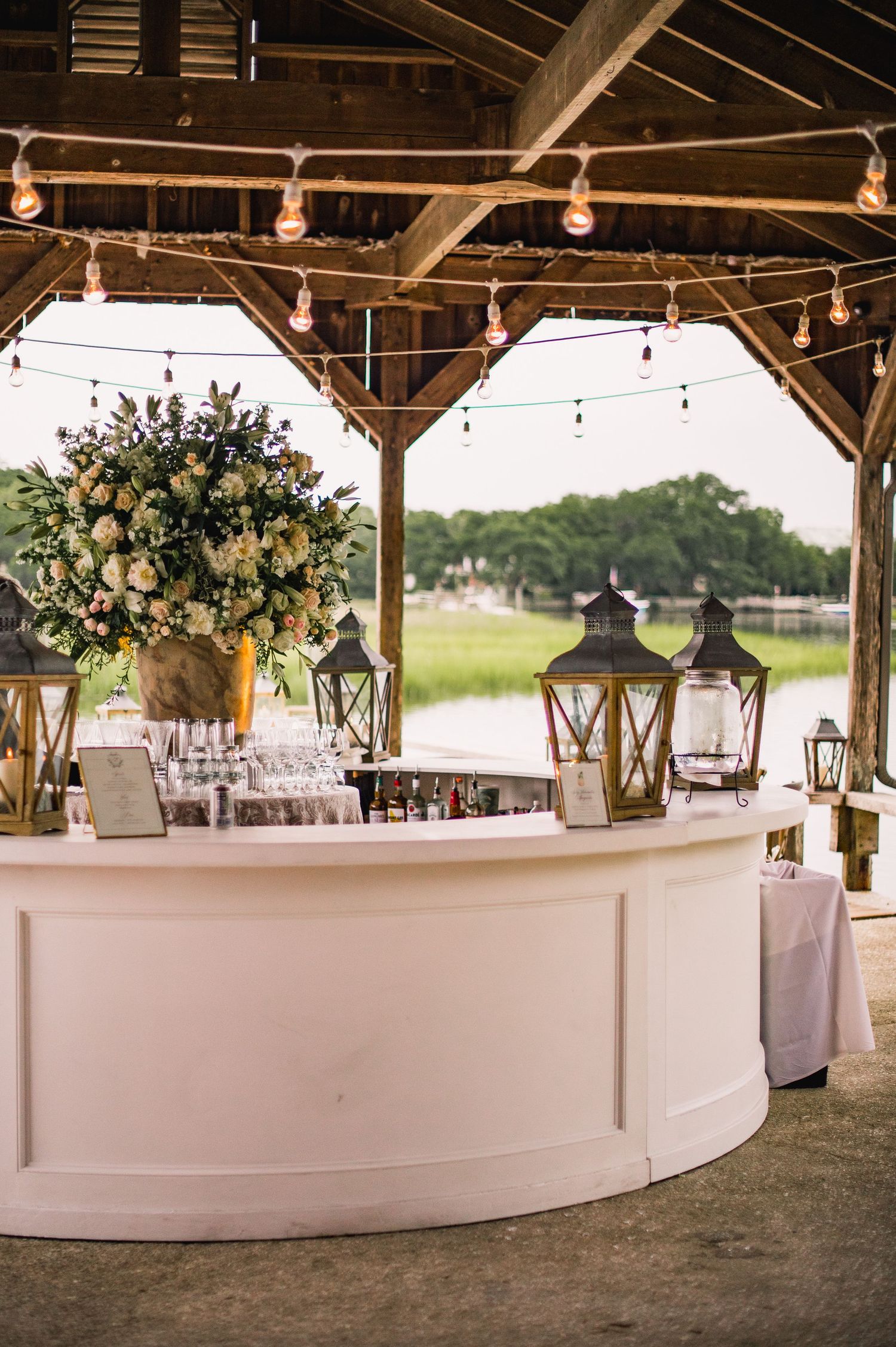 Curved white bar setup under rustic wooden pavilion with string lights and floral arrangements for outdoor event.