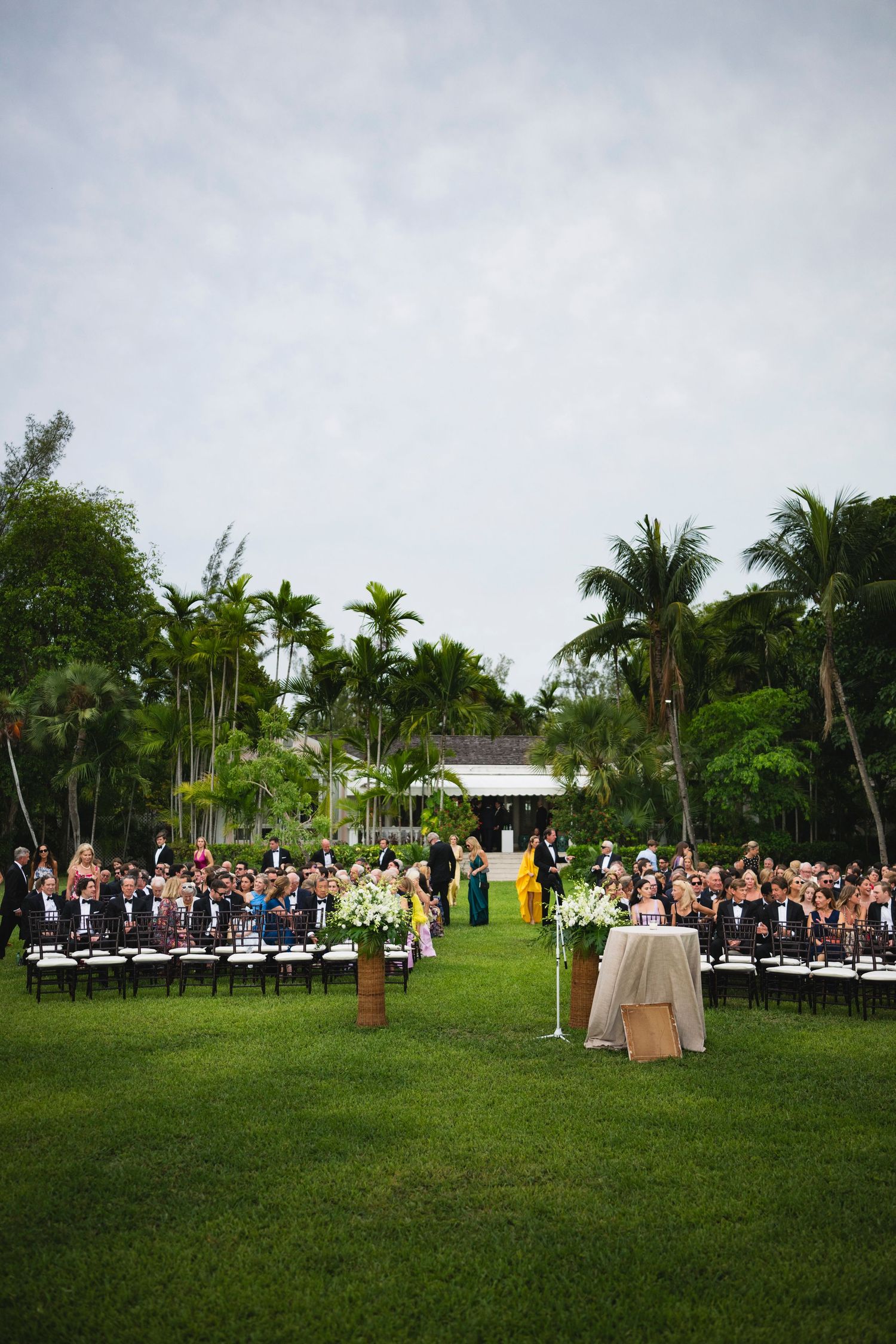 Outdoor wedding ceremony setup with rows of chairs on a lush green lawn surrounded by tropical palm trees and foliage.