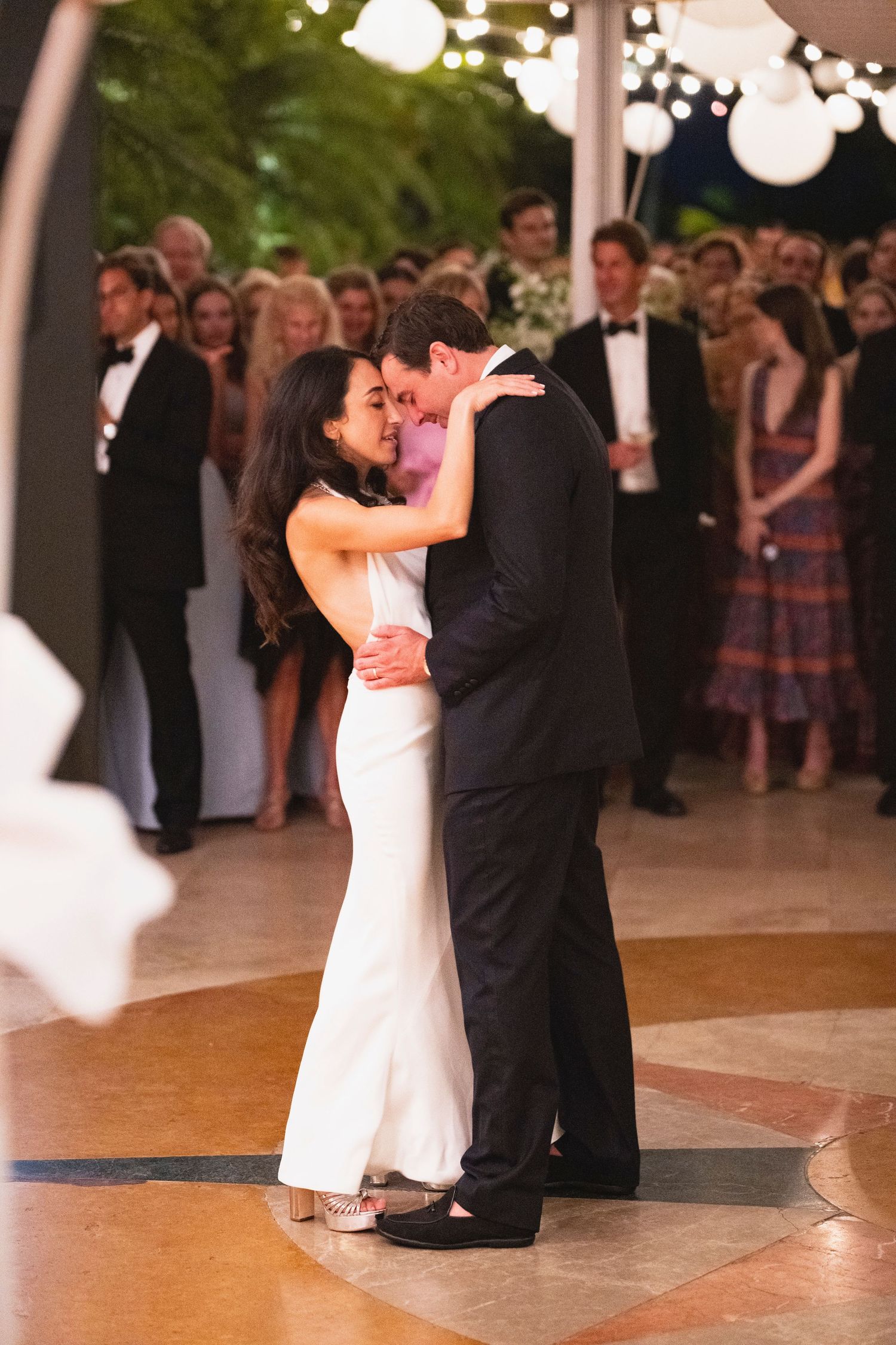 A couple shares their first dance at a wedding reception while guests look on from the background.