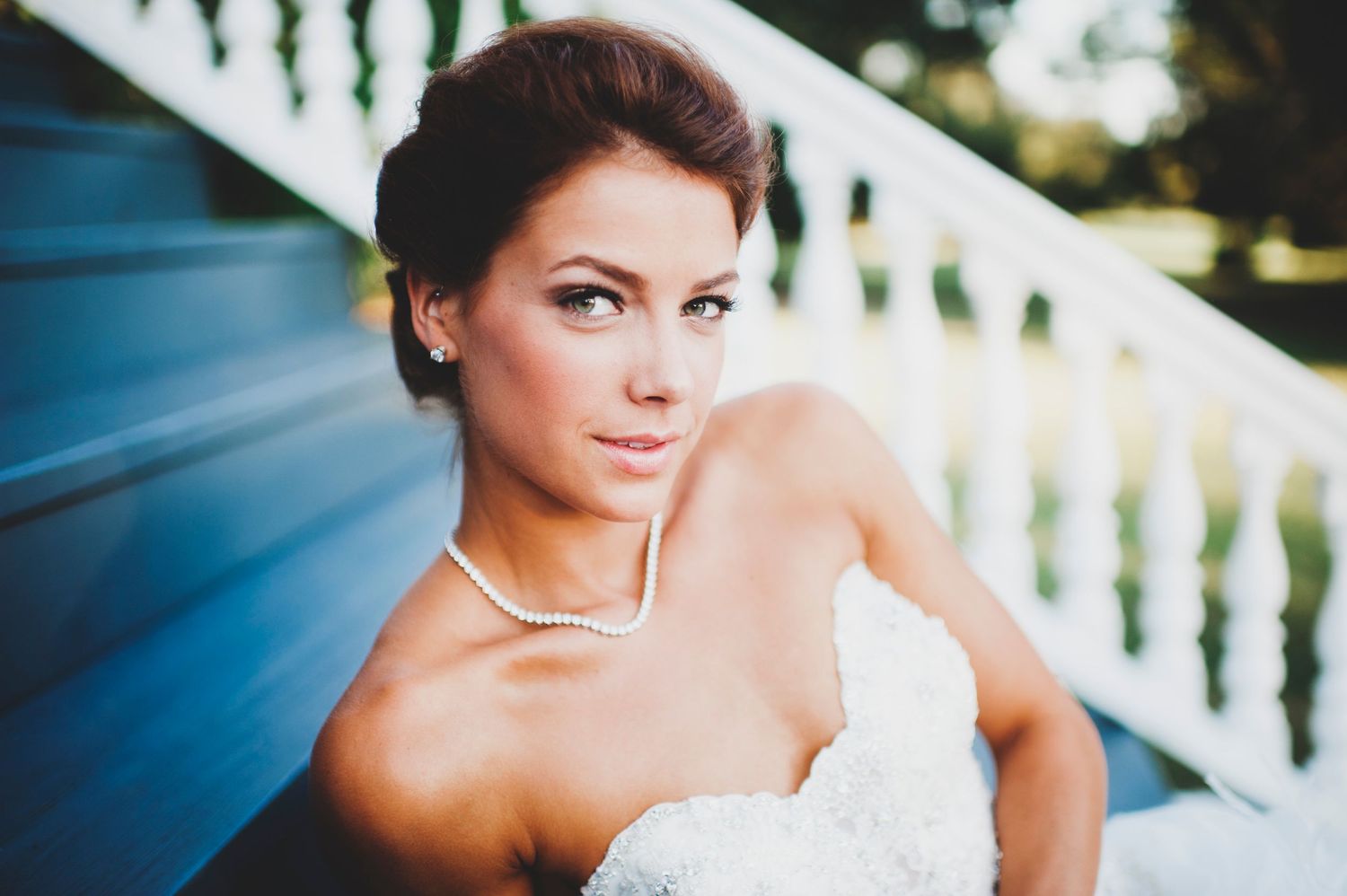 A bride in a white strapless wedding dress poses gracefully on a white balustrade in outdoor garden setting.
