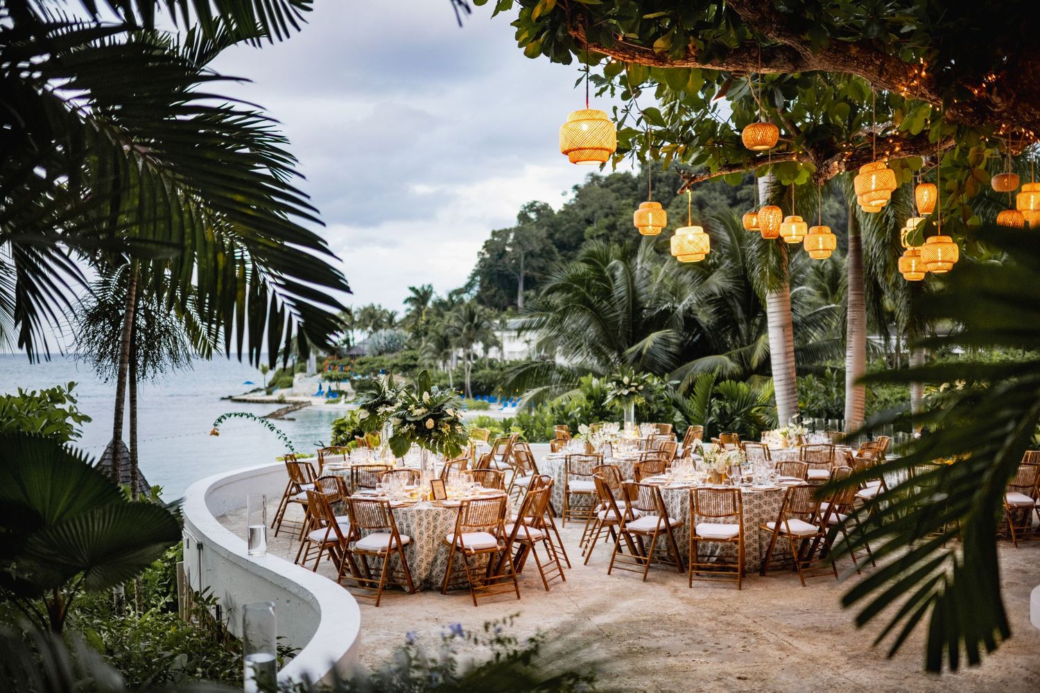 Elegant outdoor wedding reception setup with round tables, lanterns, and palm trees at dusk.