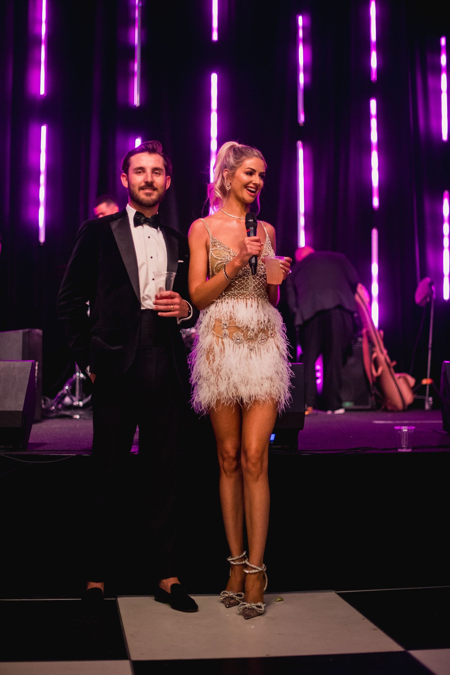 Couple in formal attire stands on stage with purple neon lighting in background at an evening event.