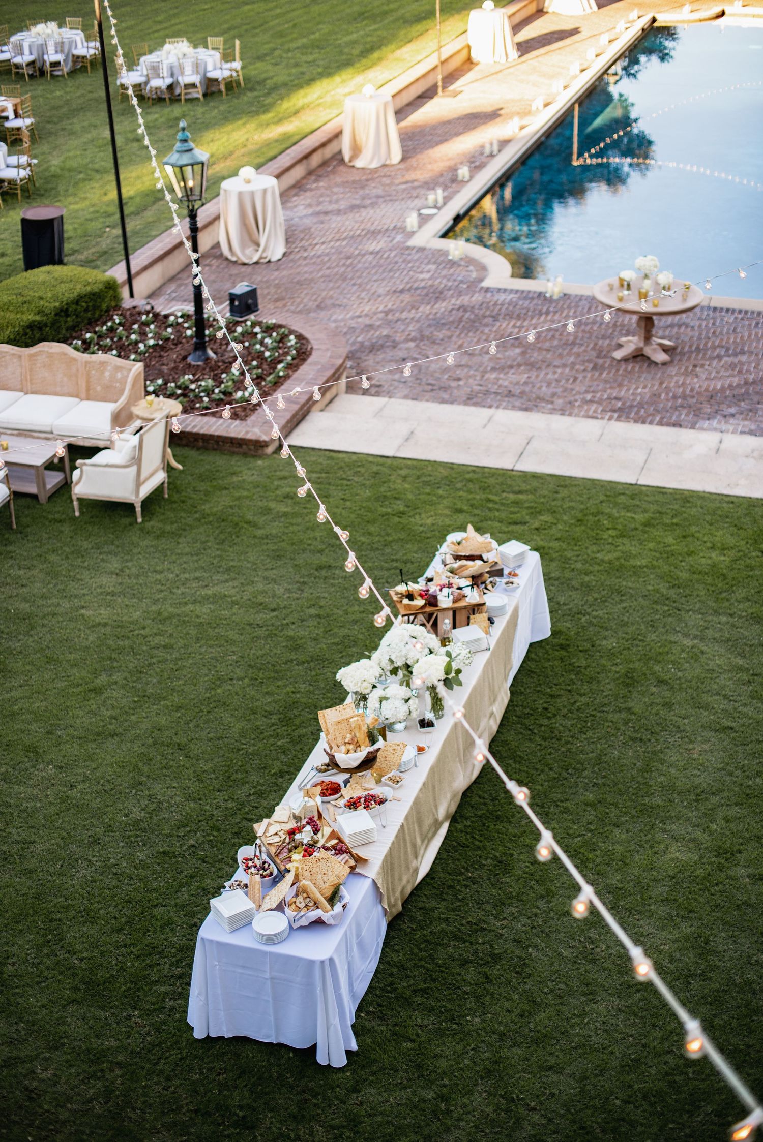 Outdoor wedding reception setup with white buffet tables and string lights on a manicured lawn at dusk.