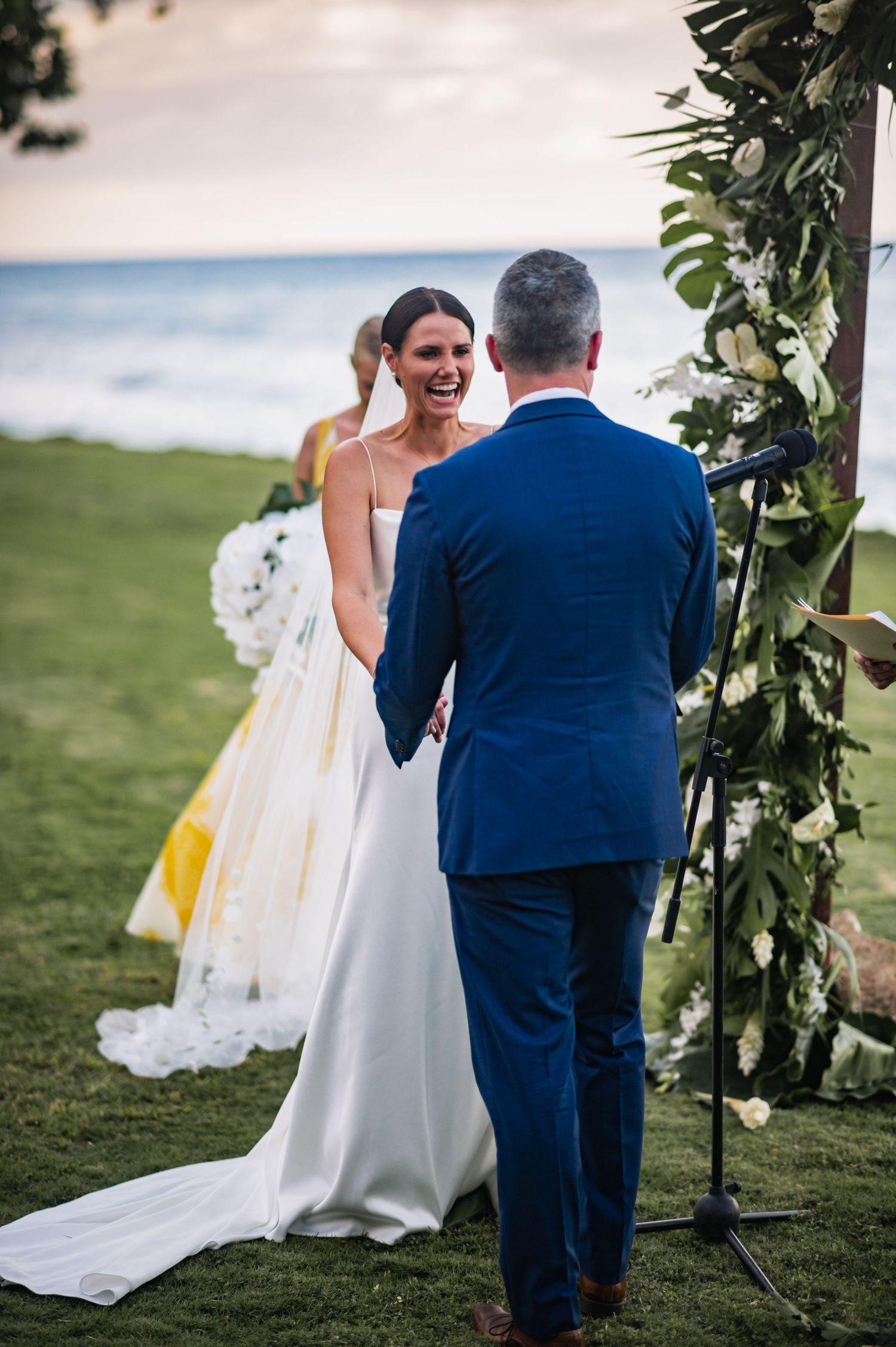 A bride in white dress and groom in navy suit share a candid moment on a grassy hillside during their wedding day.