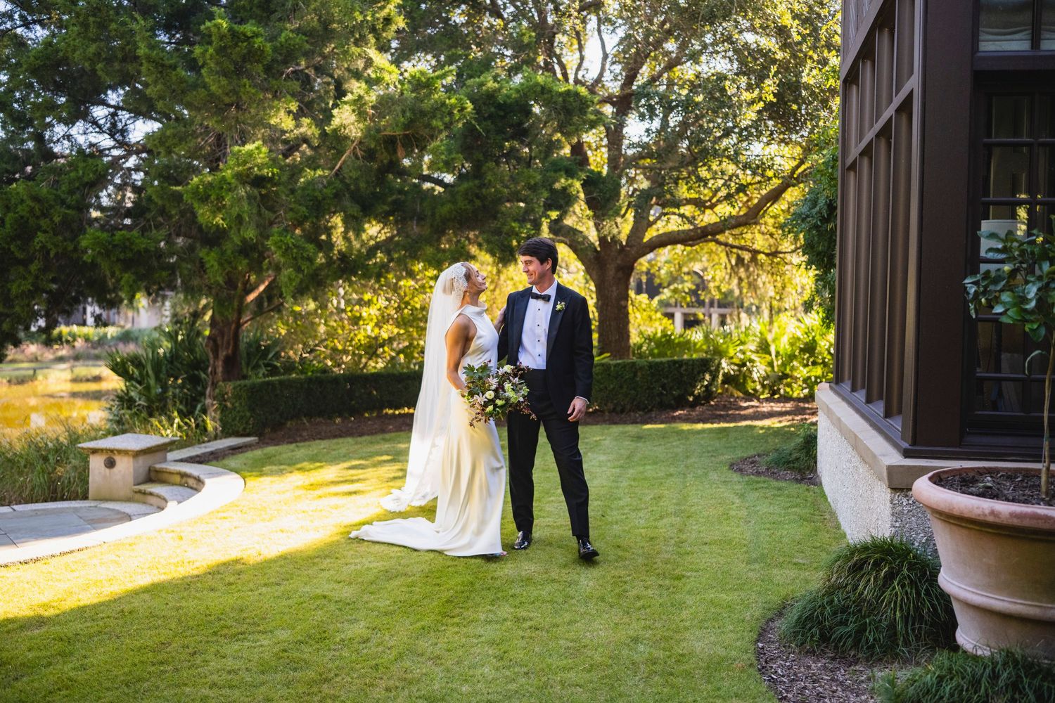 A couple in wedding attire stands together on a lush green lawn during golden hour at an outdoor ceremony.