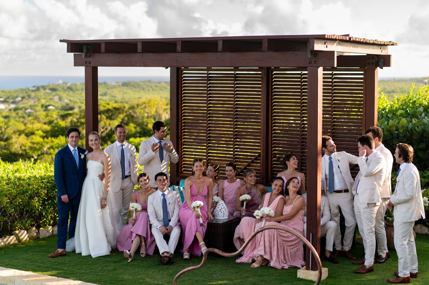 Wedding party in pink dresses and white suits posing outdoors at a tropical venue.