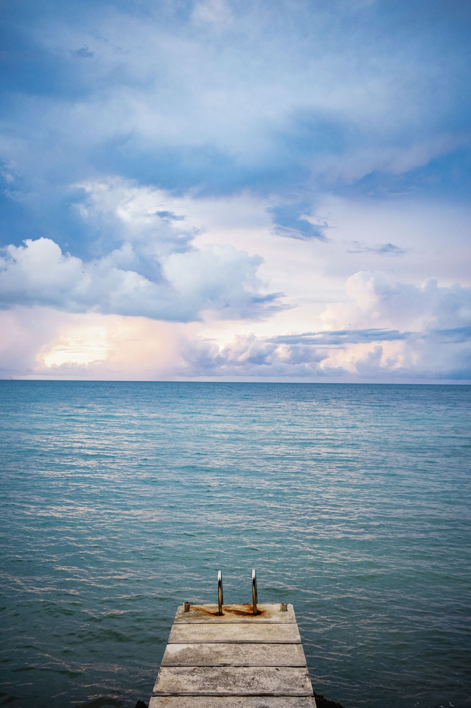 Wooden pier at Round Hill resort Jamaica extends into calm turquoise waters under blue skies with puffy white clouds.