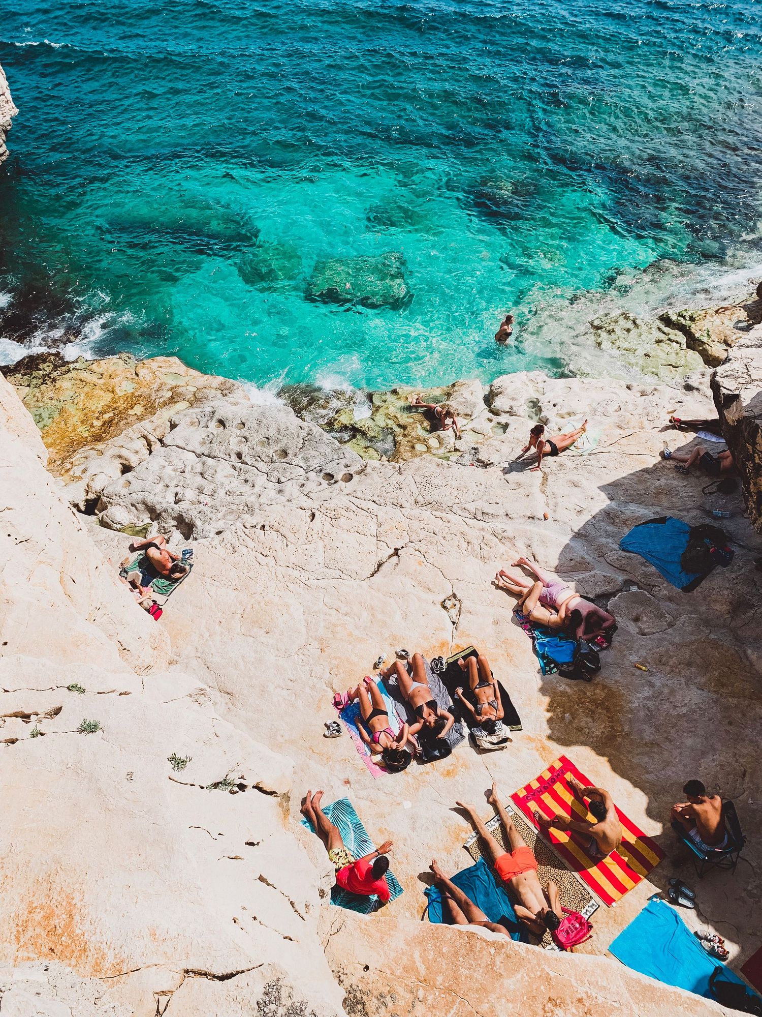Aerial view of beachgoers relaxing on rocky limestone cliffs beside vibrant turquoise Mediterranean waters.