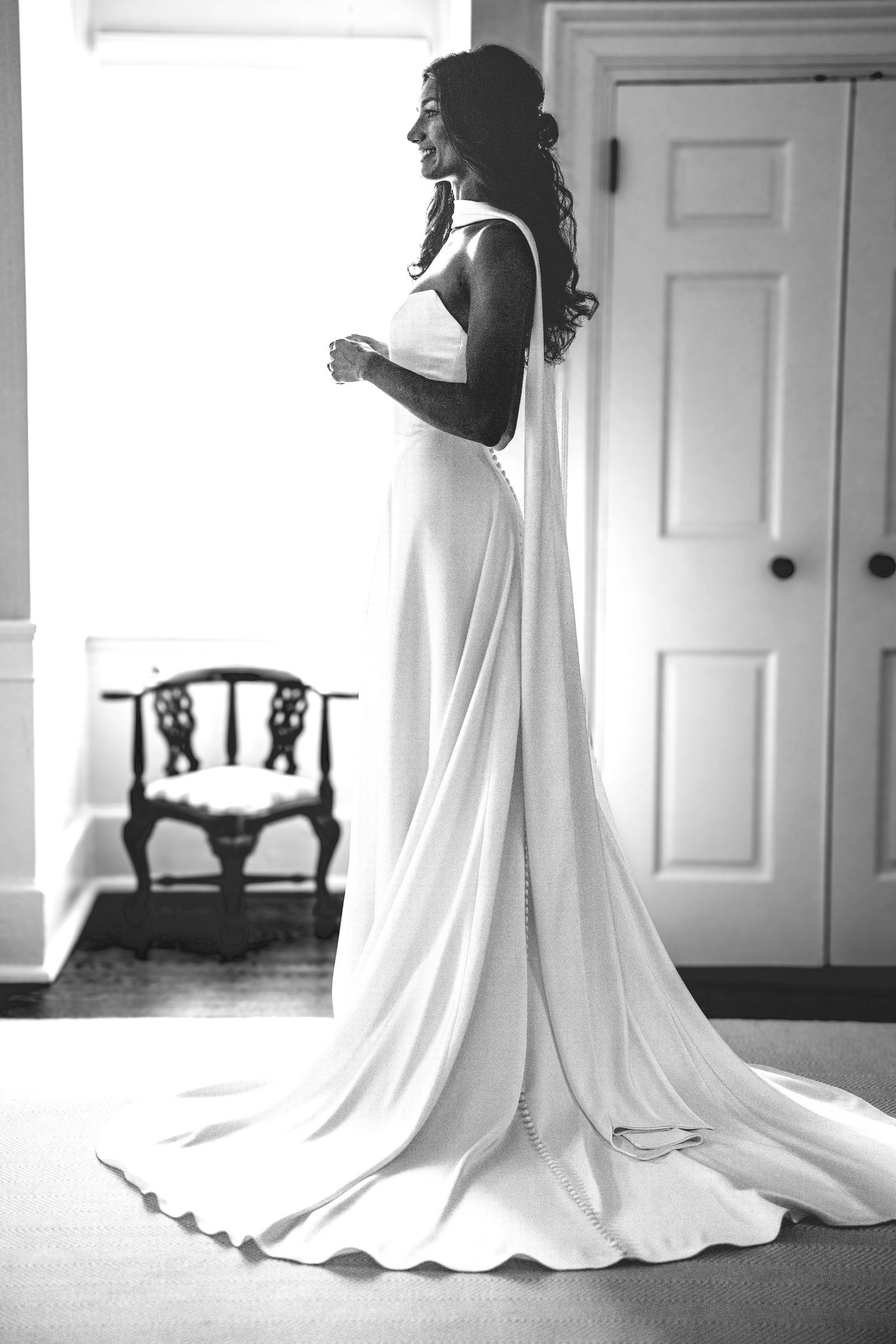 A black and white photograph shows an elegant bride in a flowing white gown with a dramatic train in a sunlit room.