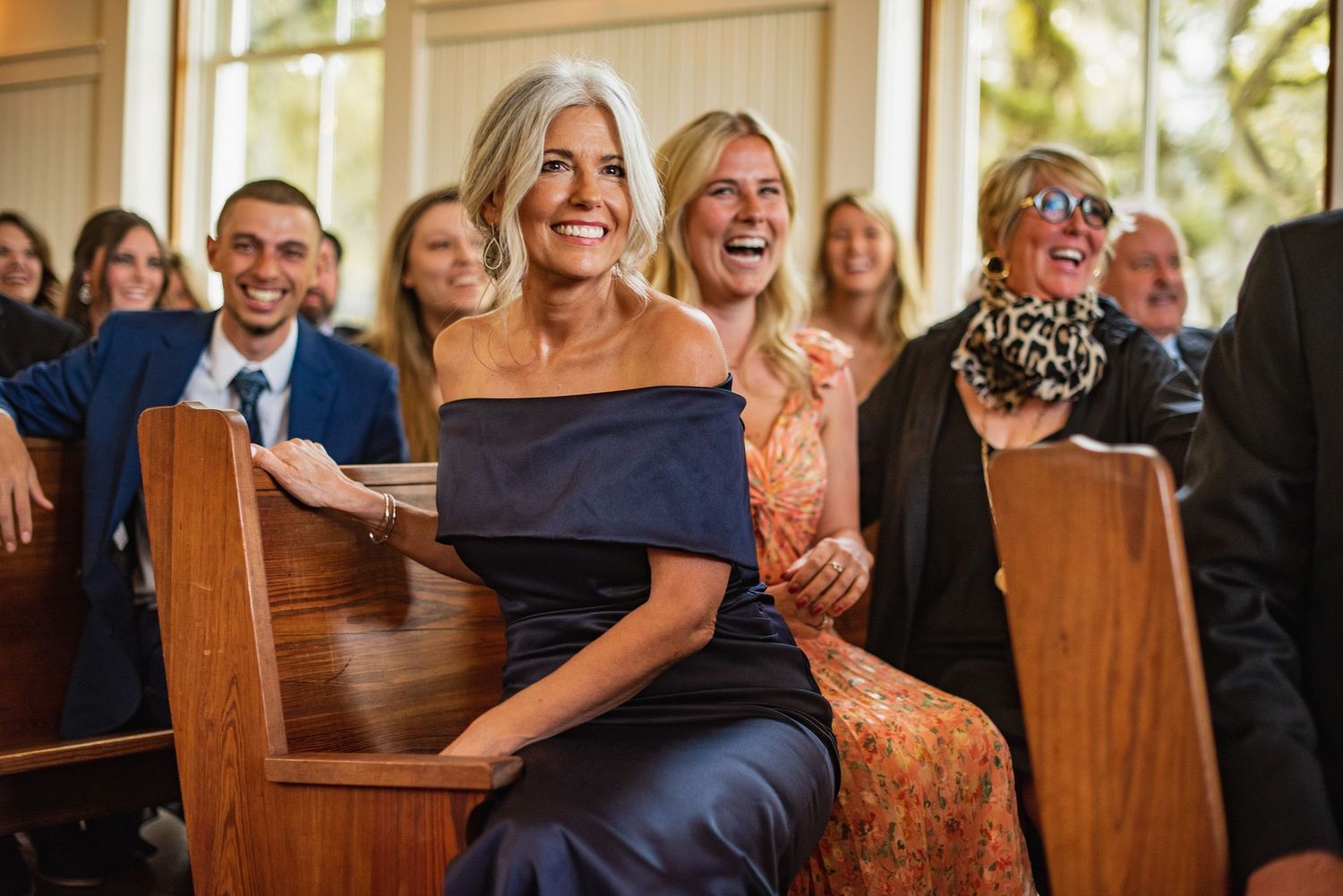 Wedding guests laugh together while seated in wooden pews during a joyful ceremony celebration.