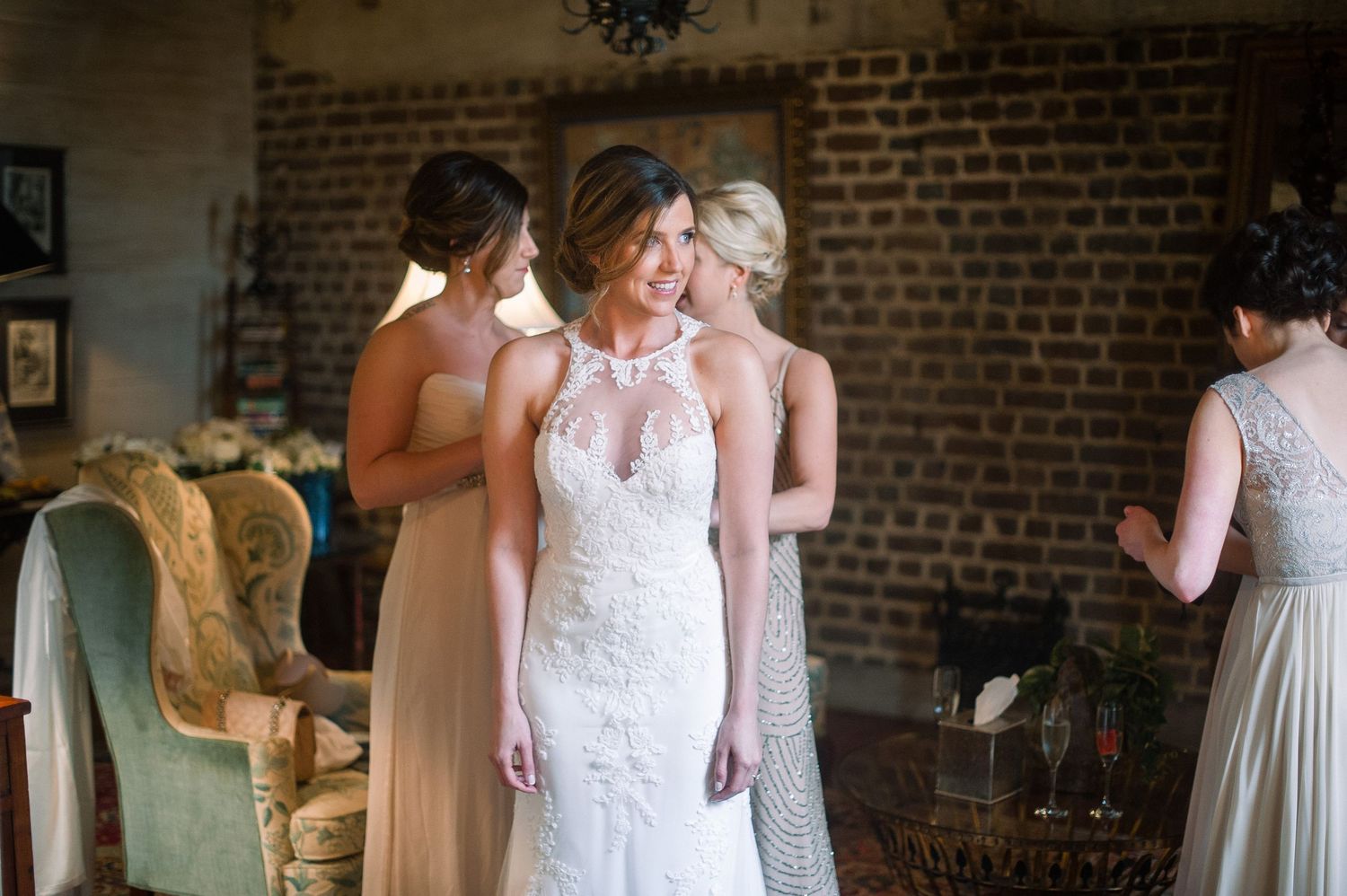 A bride in an elegant lace wedding gown stands in a rustic brick-walled room with vintage furniture.