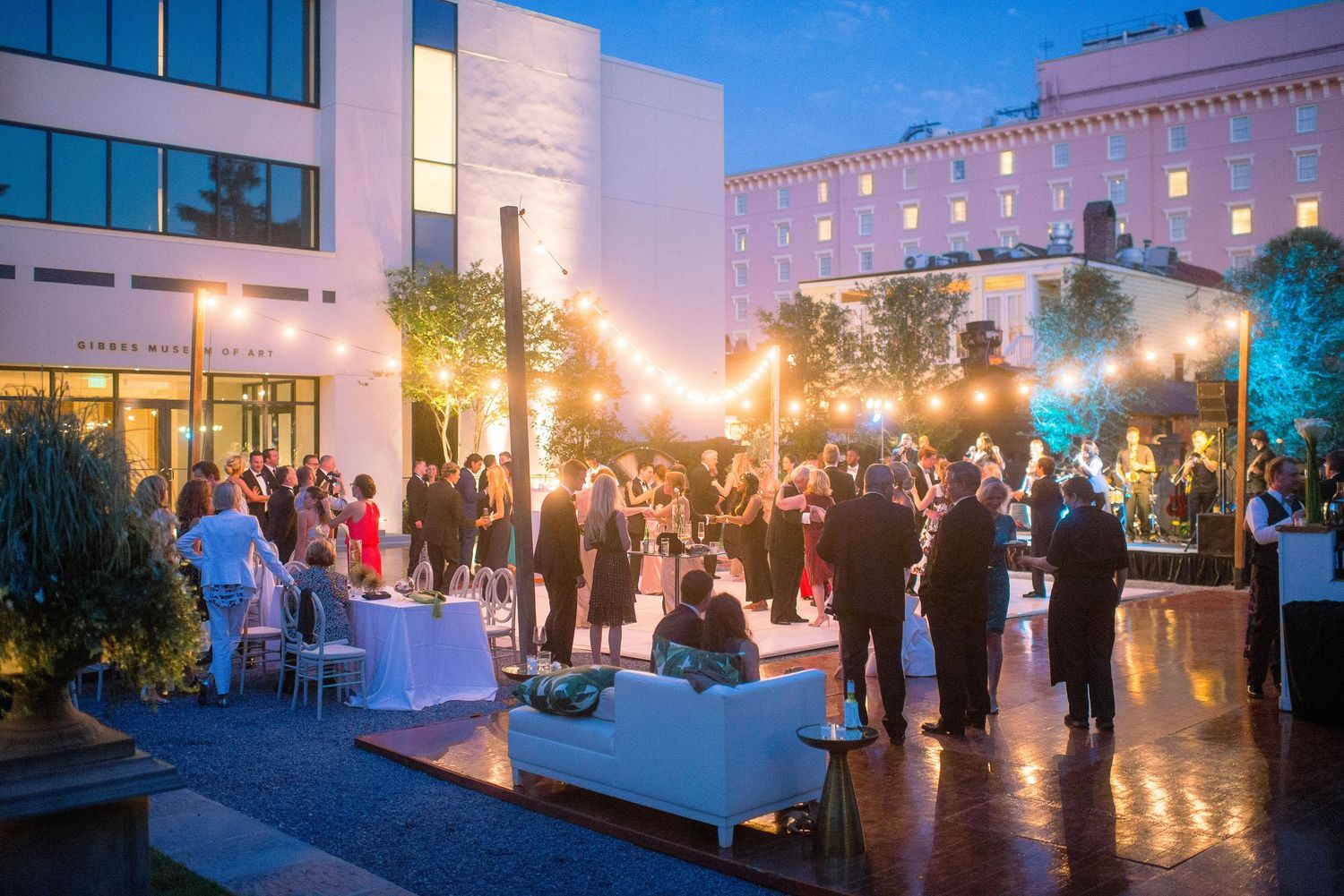 Elegant outdoor evening event with guests mingling on a terrace illuminated by warm lighting and glowing lanterns.
