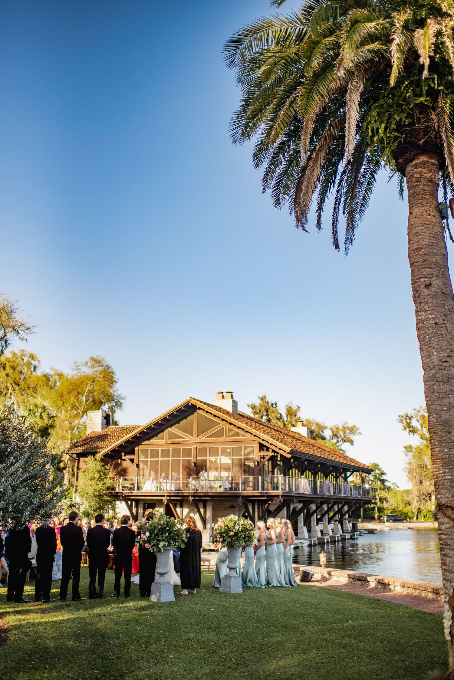 Wedding ceremony taking place at sunset on a lush lawn with palm trees and a rustic venue building in the background.