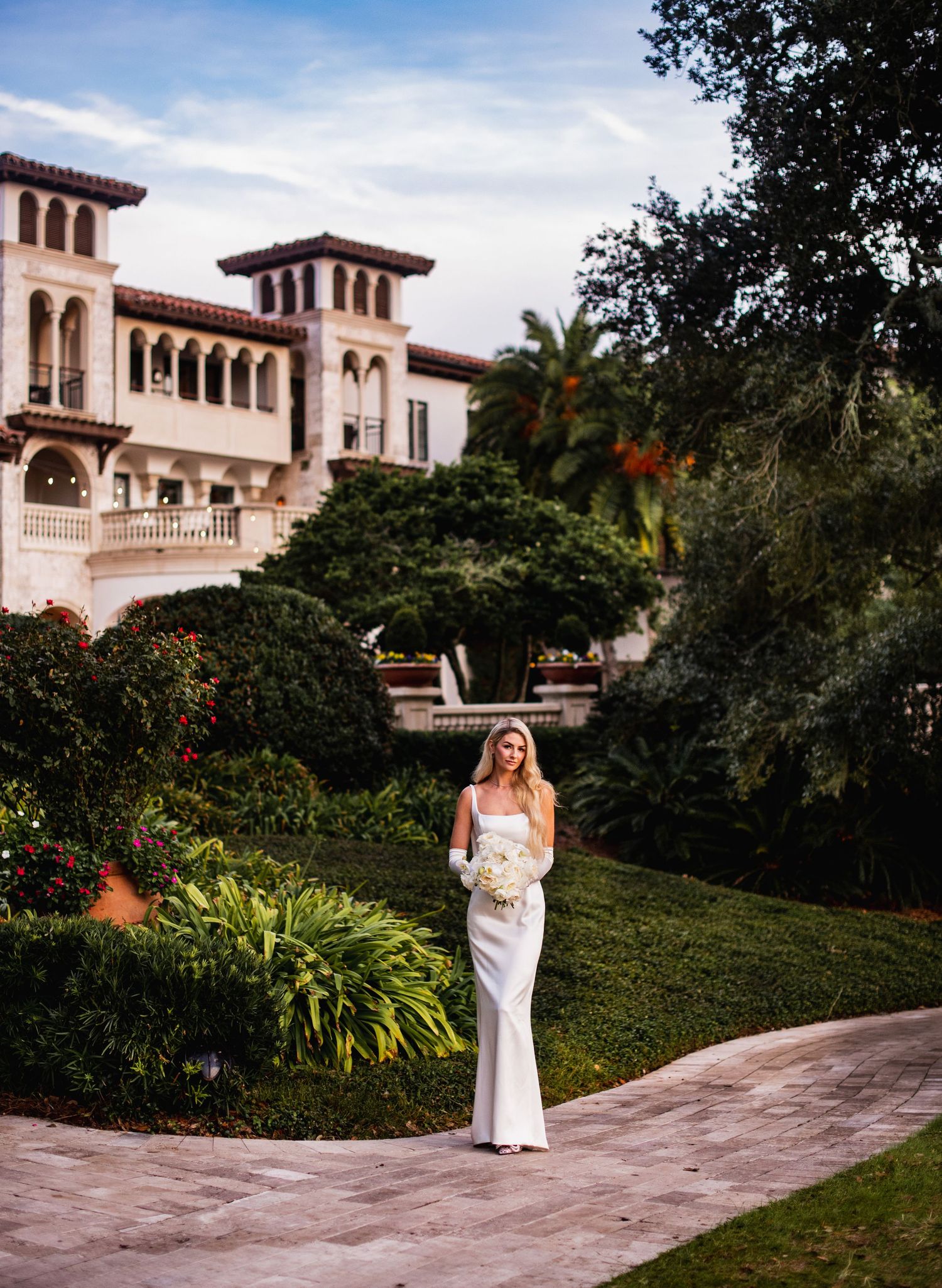 Bride in elegant white gown poses in front of Mediterranean-style mansion with lush gardens.