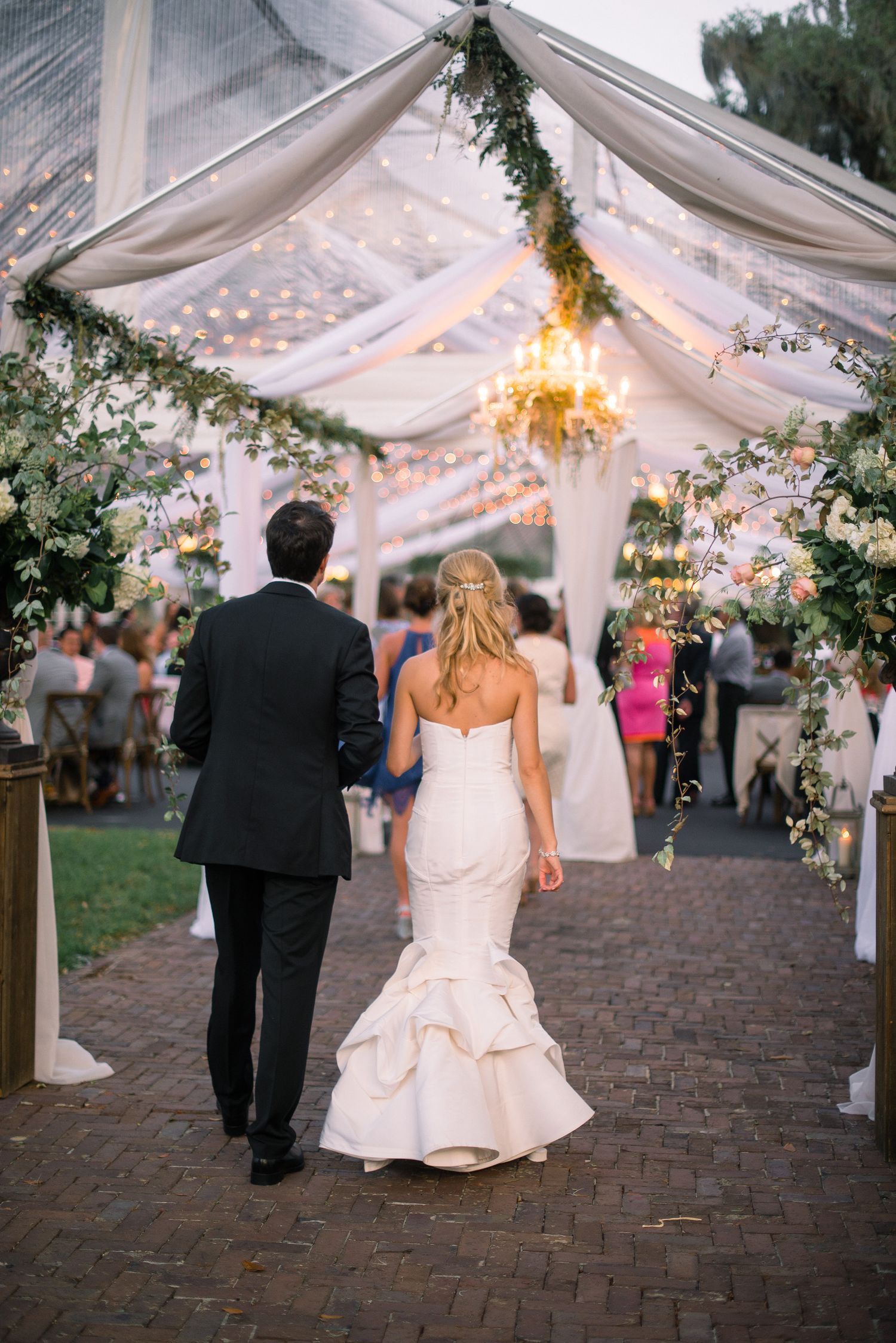 A couple walks under an elegant outdoor wedding tent decorated with twinkling lights, white drapes and greenery garlands.