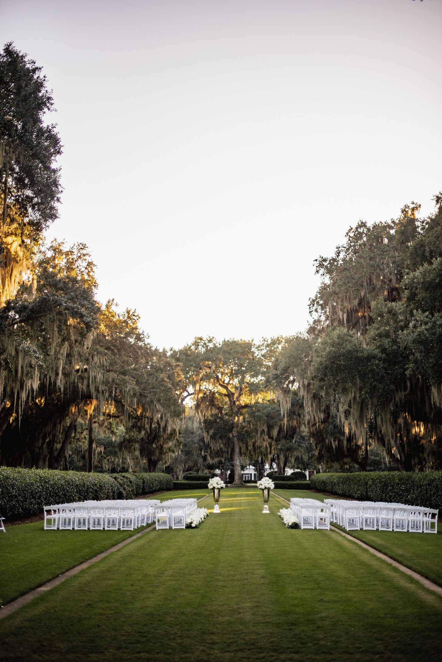 White chairs line a grassy outdoor ceremony space framed by Spanish moss-draped oak trees at sunset.