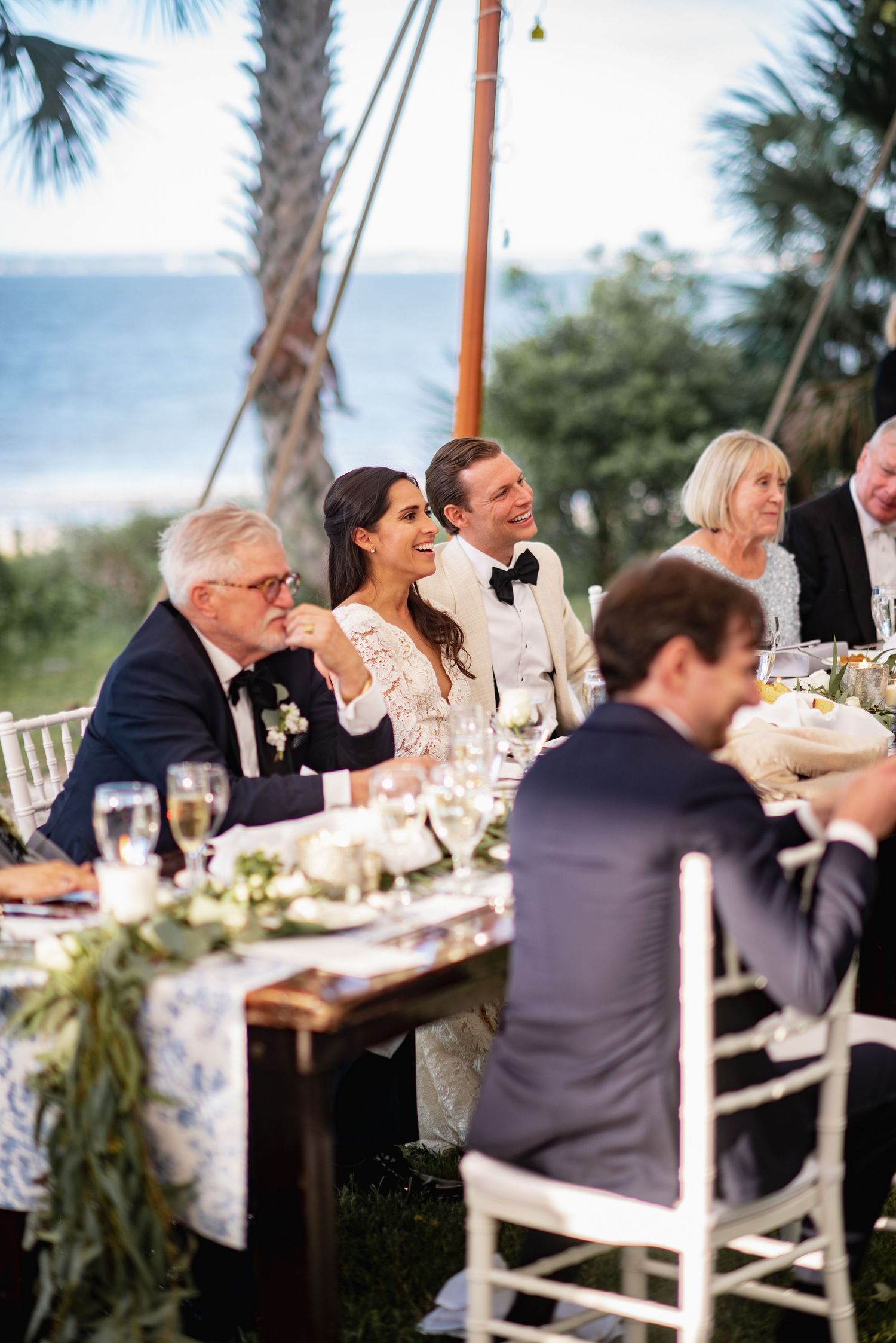 Wedding guests seated at an elegant outdoor reception table under palm trees share conversations over dinner and drinks.