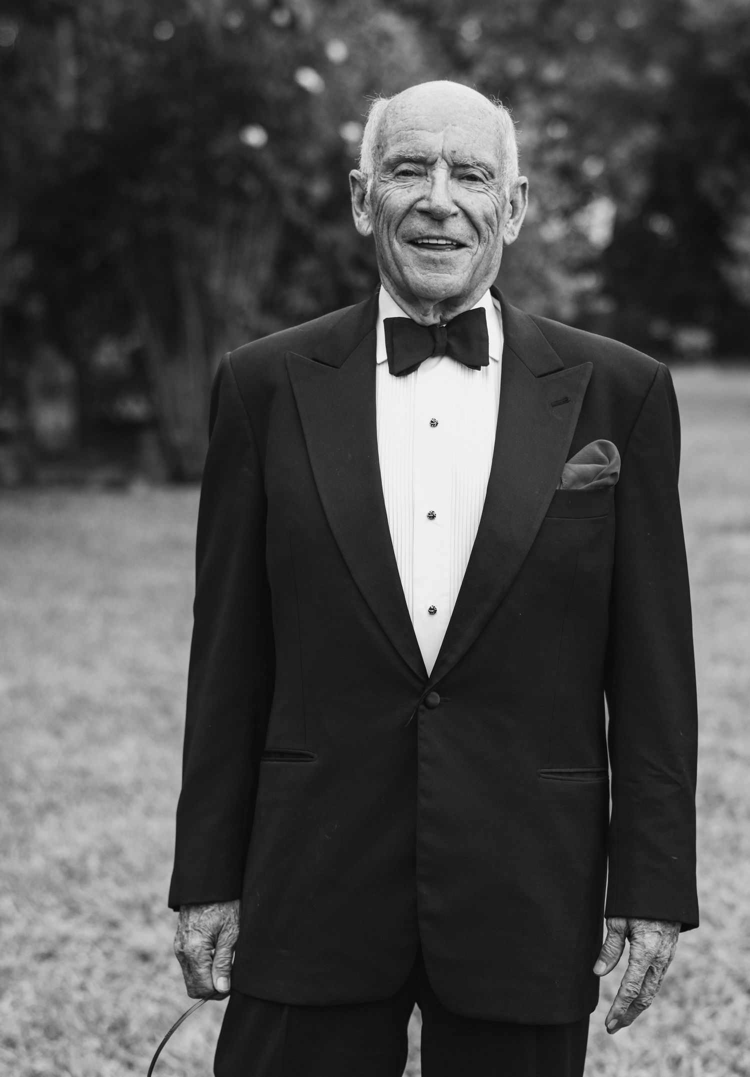 A black and white portrait of someone in a formal tuxedo with bow tie smiling outdoors.